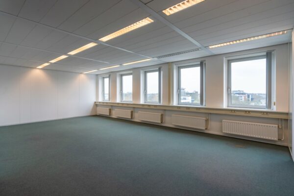 Leeg kantoor met blauwe vloerbedekking, systeemplafond en vijf ramen aan de buitenmuur in het gebouw aan de Printerweg.