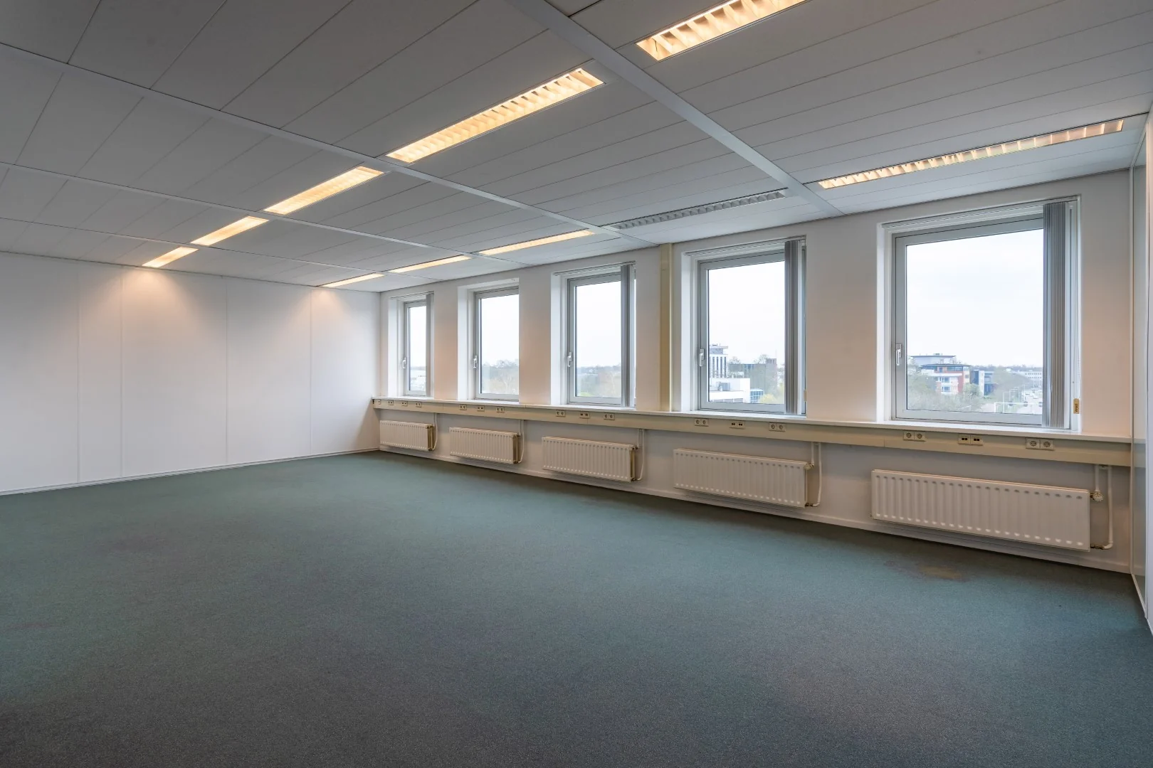 Leeg kantoor met blauwe vloerbedekking, systeemplafond en vijf ramen aan de buitenmuur in het gebouw aan de Printerweg.