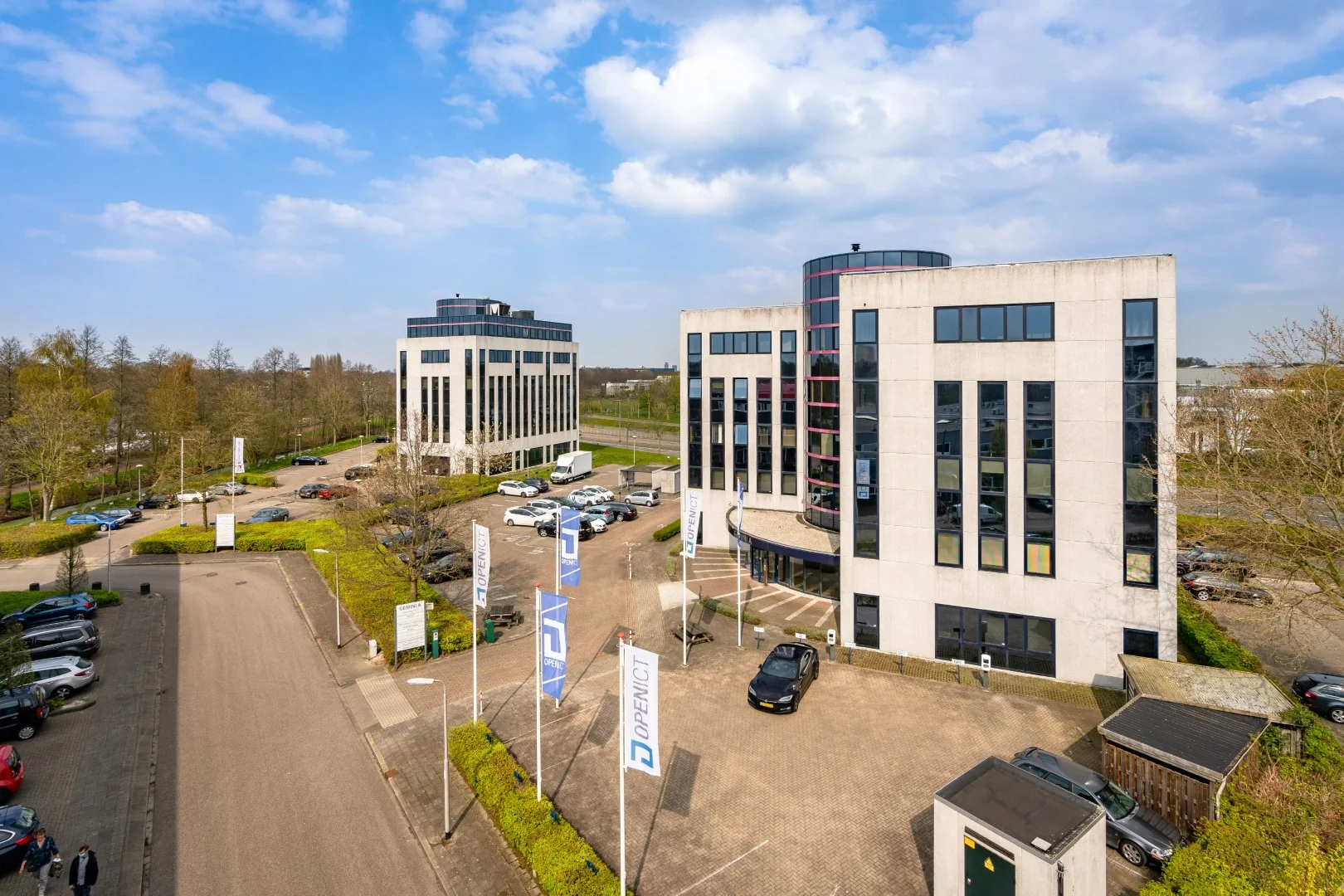 Twee moderne kantoorgebouwen aan de Printerweg met parkeerplaatsen en vlaggen van OpenICT voor de ingang.