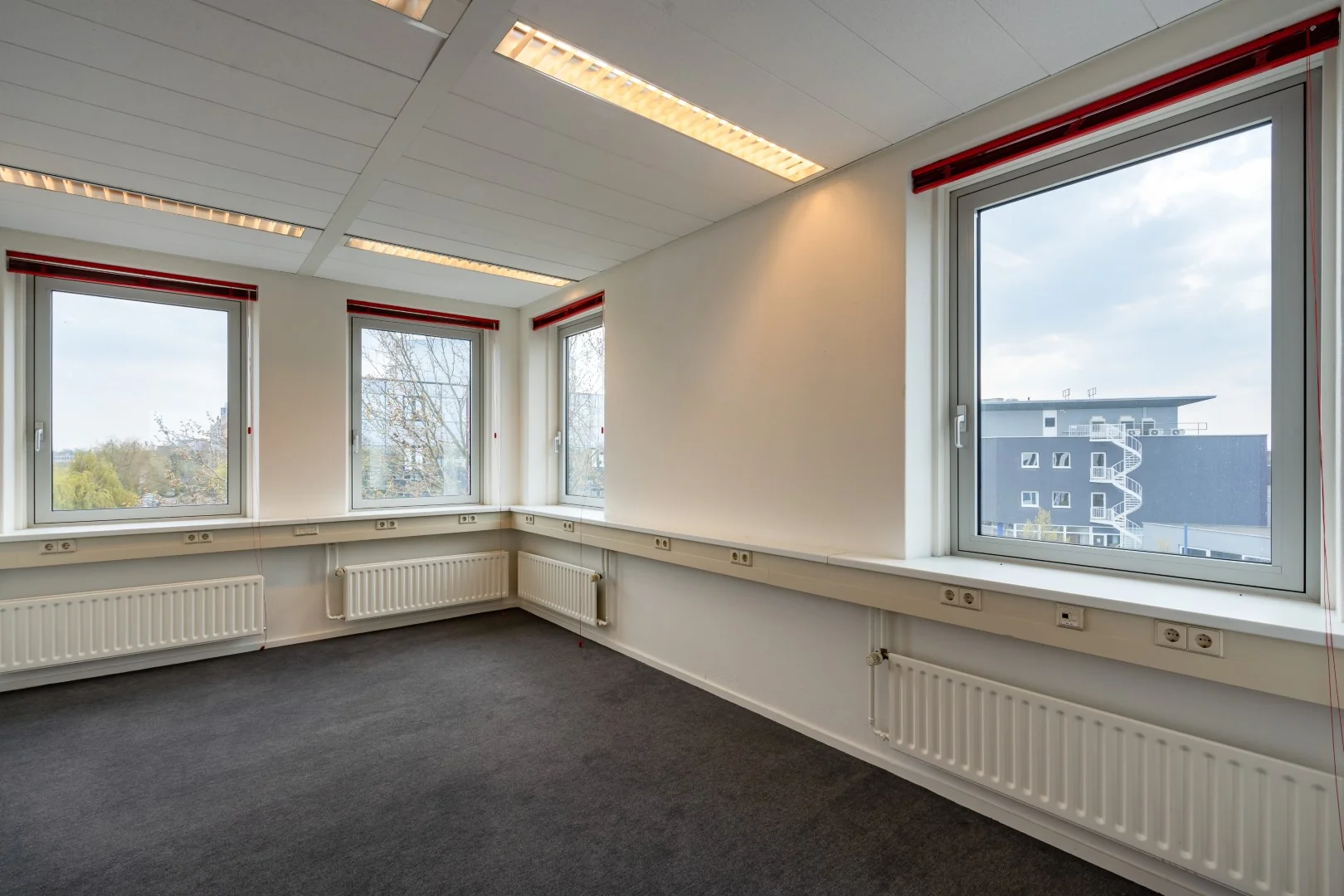 Lichte, lege kantoorruimte aan de Printerweg met vier grote ramen en zicht op een modern gebouw met buitentrap.