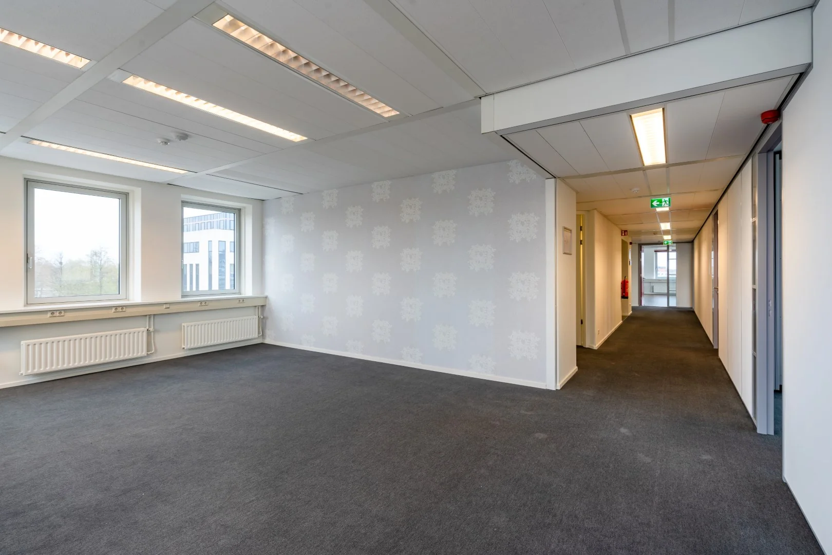 Leeg kantoorruimte met grijs tapijt, grote ramen en een gang met nooduitgang in een gebouw aan de Printerweg.