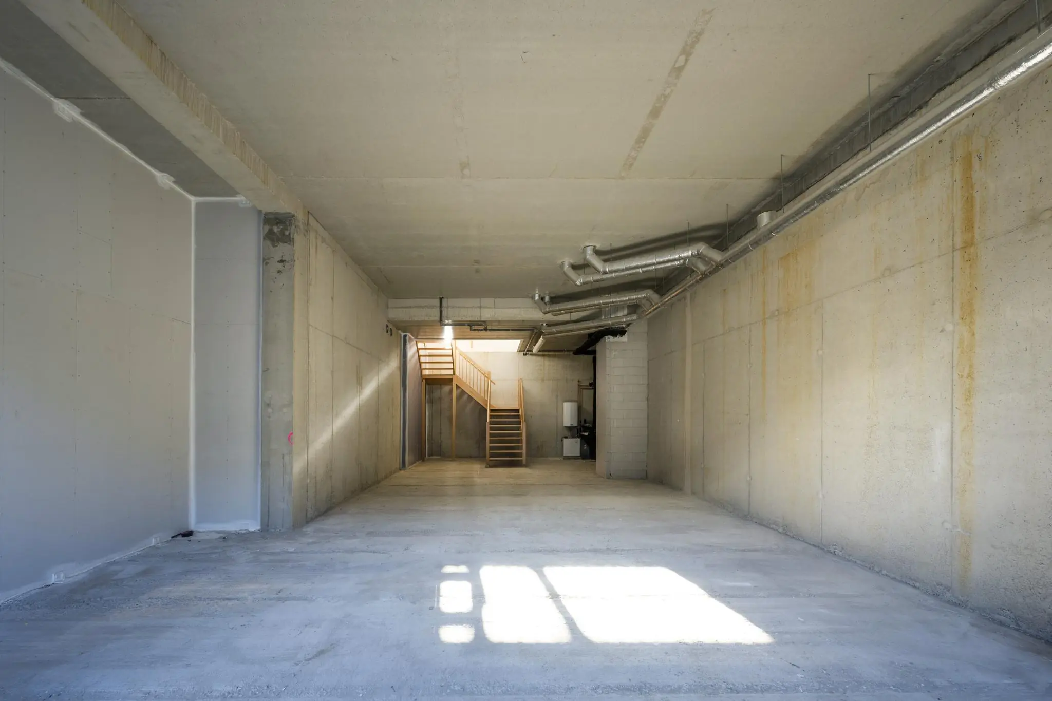 Lege, industriële ruimte aan de Oostenburgermiddenstraat met betonnen muren, zichtbare ventilatiebuizen en een houten trap achterin.