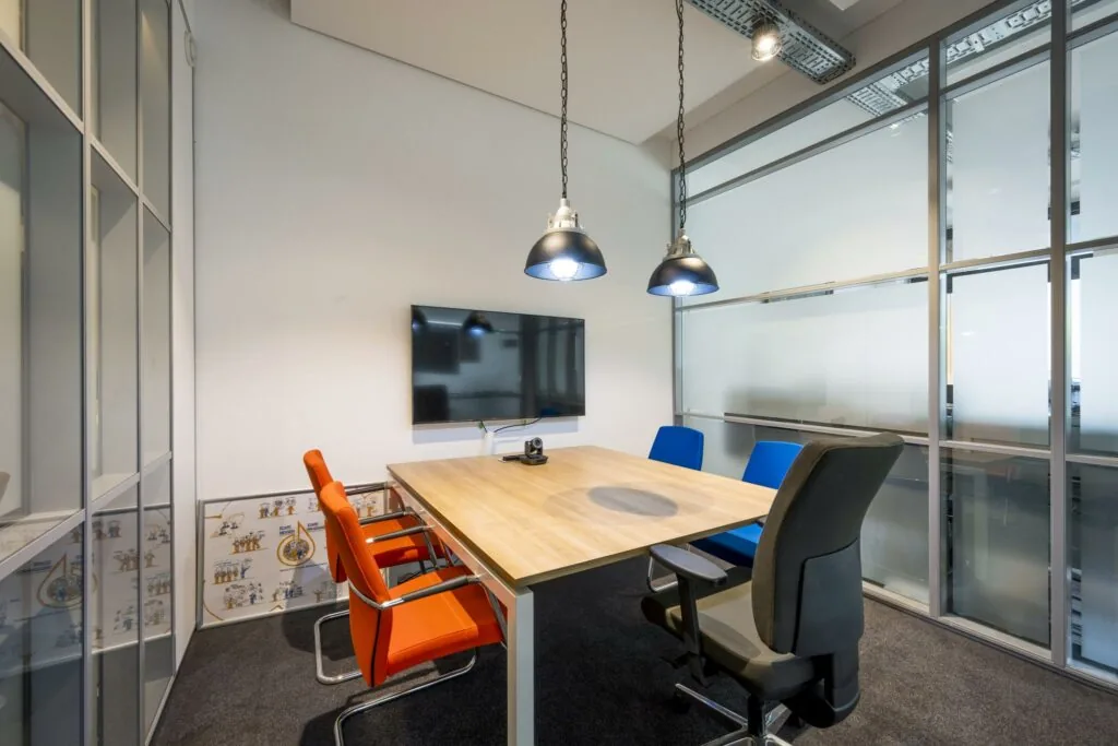 Vergaderruimte op de Asterweg met een moderne inrichting, glazen wanden, een houten tafel, kleurrijke stoelen en een scherm aan de muur.
