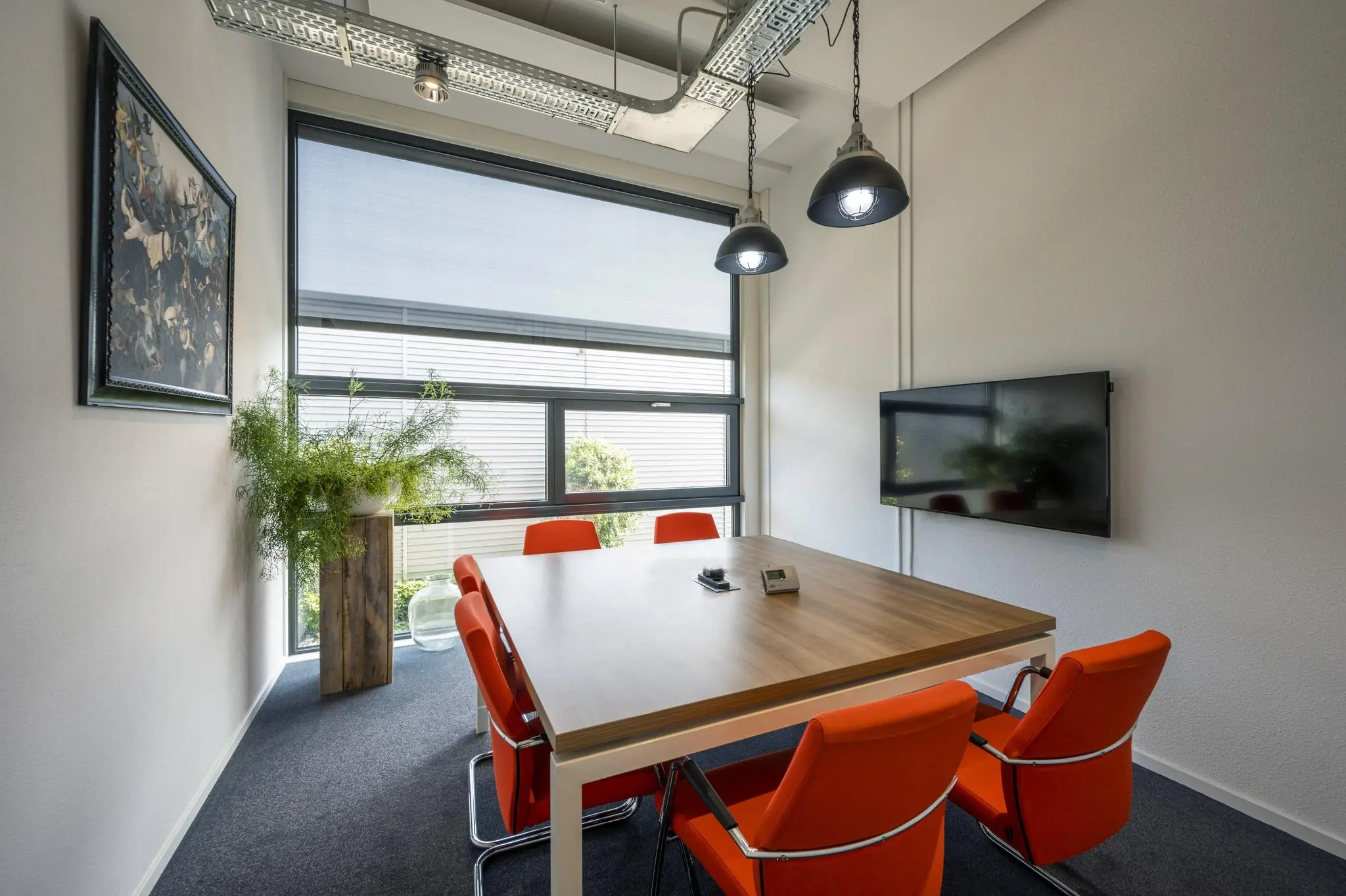 Vergaderruimte met houten tafel, feloranje stoelen, een flatscreen aan de muur en een groot raam met natuurlijk licht.