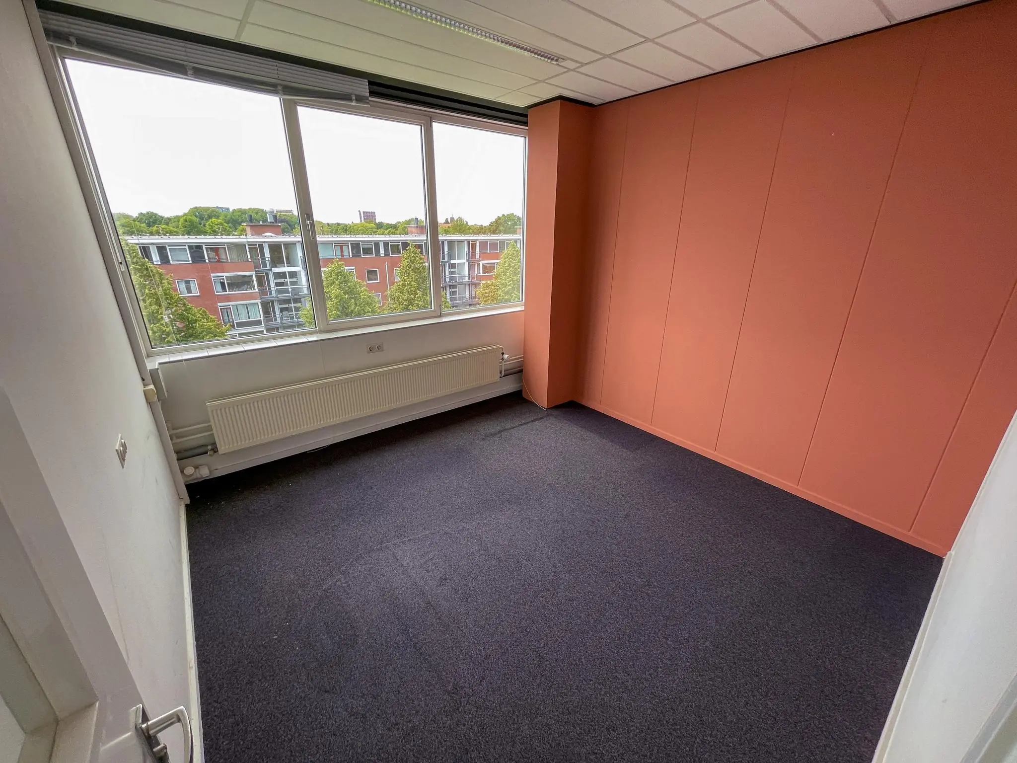 Leeg kantoor met oranje accentmuur, donker tapijt en een groot raam met uitzicht op appartementen aan de Hengeveldstraat.