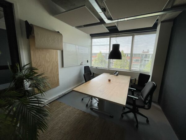 Vergaderruimte aan de Hengeveldstraat met houten tafel, vier stoelen, whiteboard en groot raam met uitzicht op een kantoorgebouw.