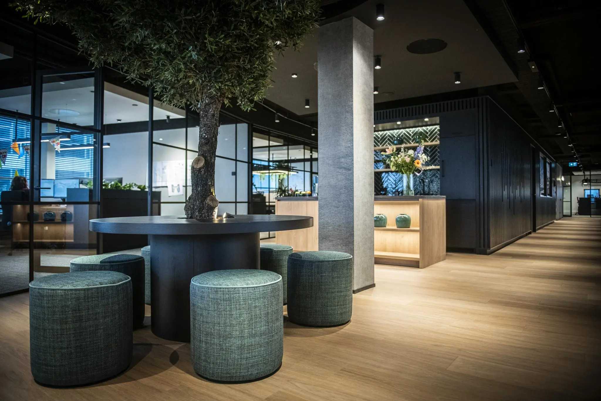 Moderne kantoorruimte in Stationspark met houten vloeren, glazen wanden, een ronde tafel met boomstam en turquoise poefjes.