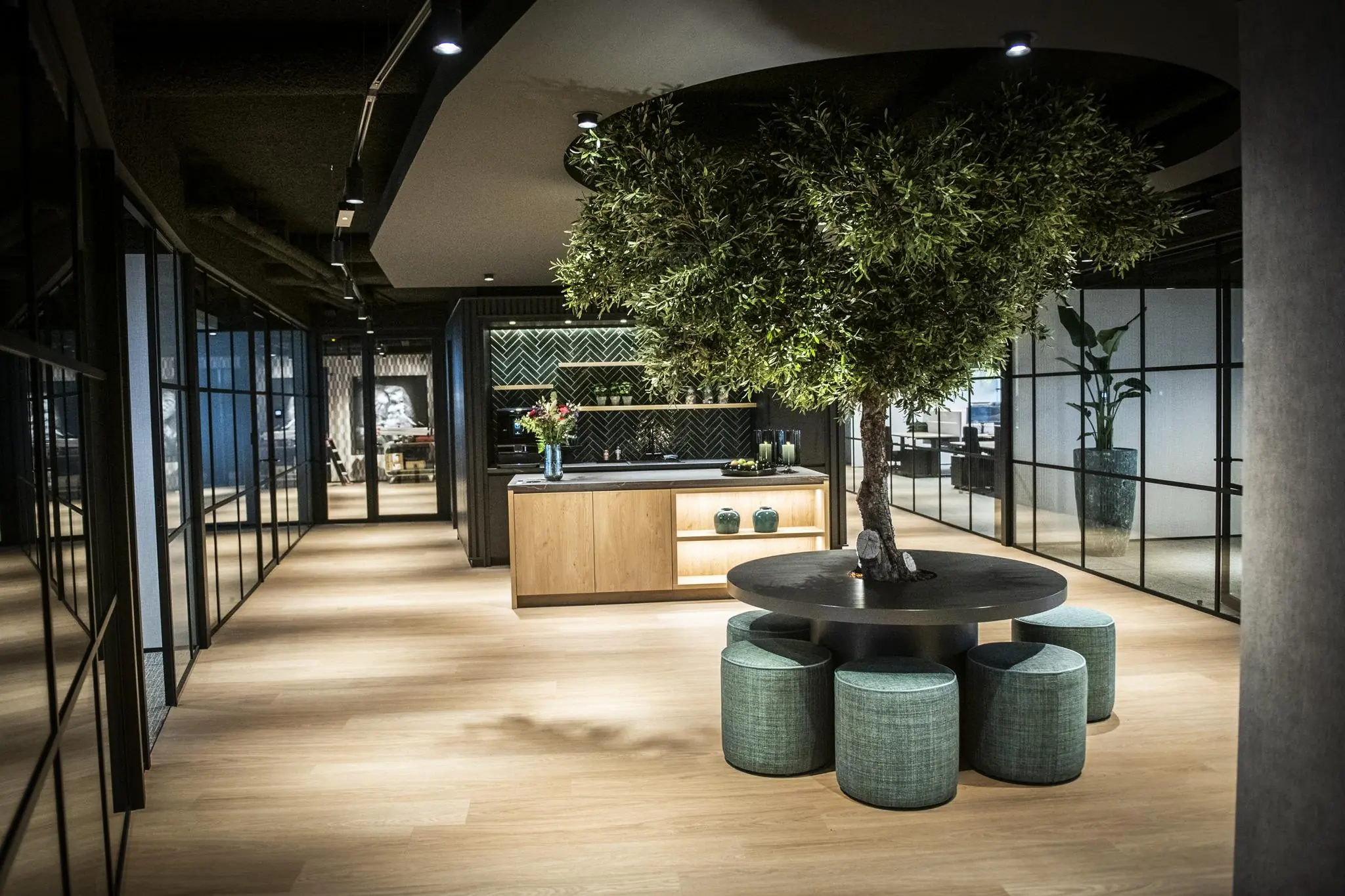 Moderne kantoorruimte in Stationspark met een ronde tafel rondom een boom, omringd door poefjes en glazen vergaderruimtes.