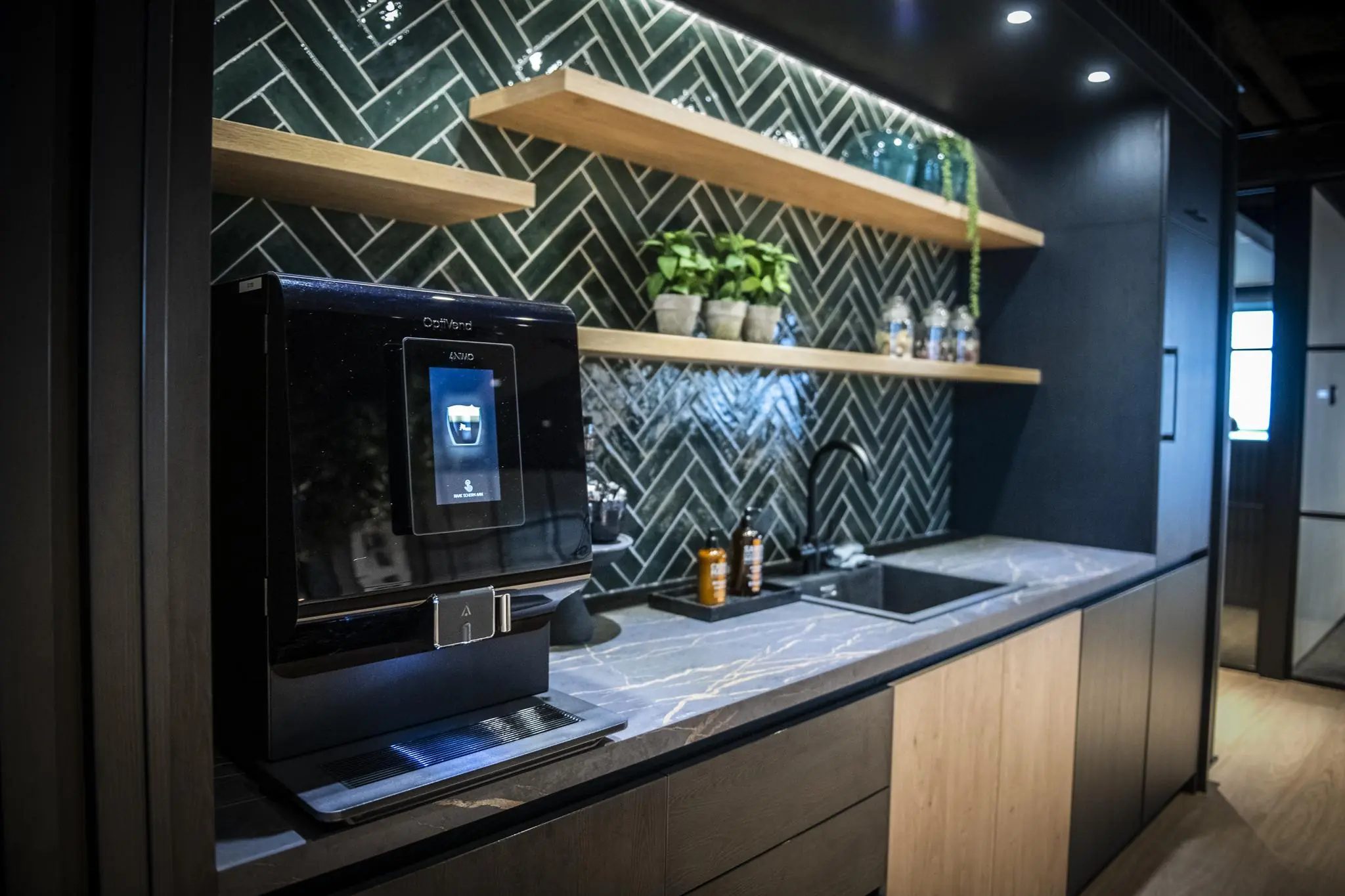 Moderne pantry met koffieautomaat, marmeren werkblad, donkergroene visgraattegels en houten wandplanken met planten en voorraadpotten.