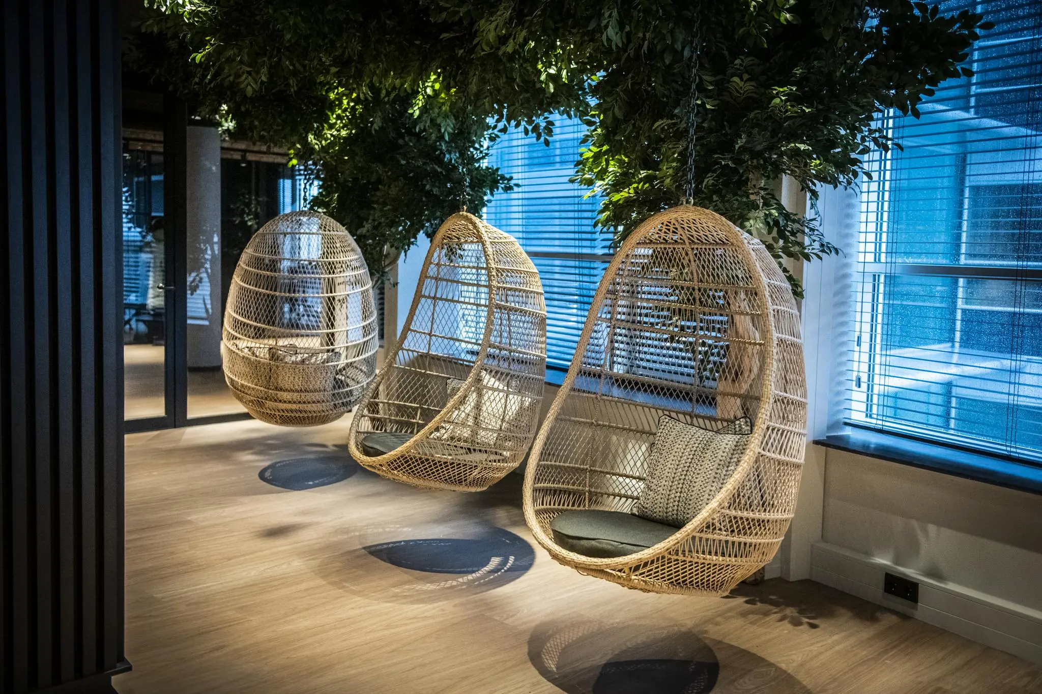 Drie rotan hangstoelen met kussens hangen aan het plafond in een modern en groen ingericht interieur van Stationspark.