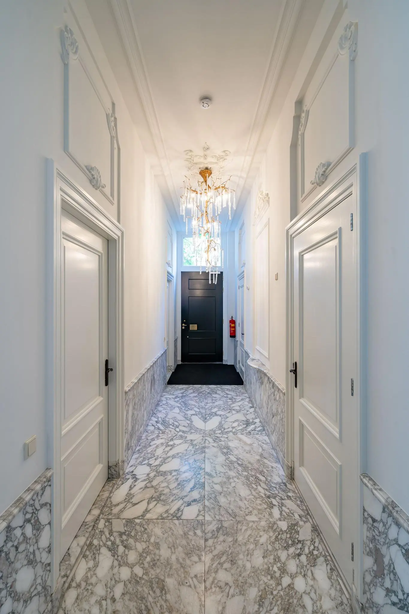 Elegante hal met marmeren vloer, witte paneeldeuren en een gouden kristallen kroonluchter aan het plafond op de Herengracht.
