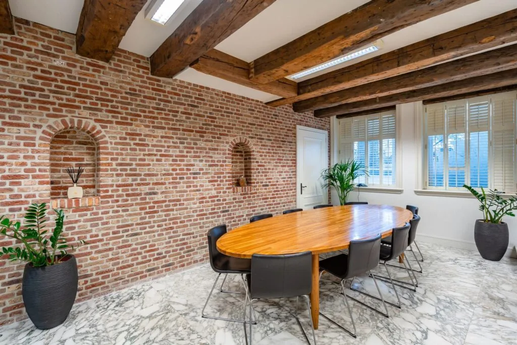 Vergaderruimte aan de Herengracht met een ovale houten tafel, bakstenen muur, houten balkenplafond en marmeren vloer.