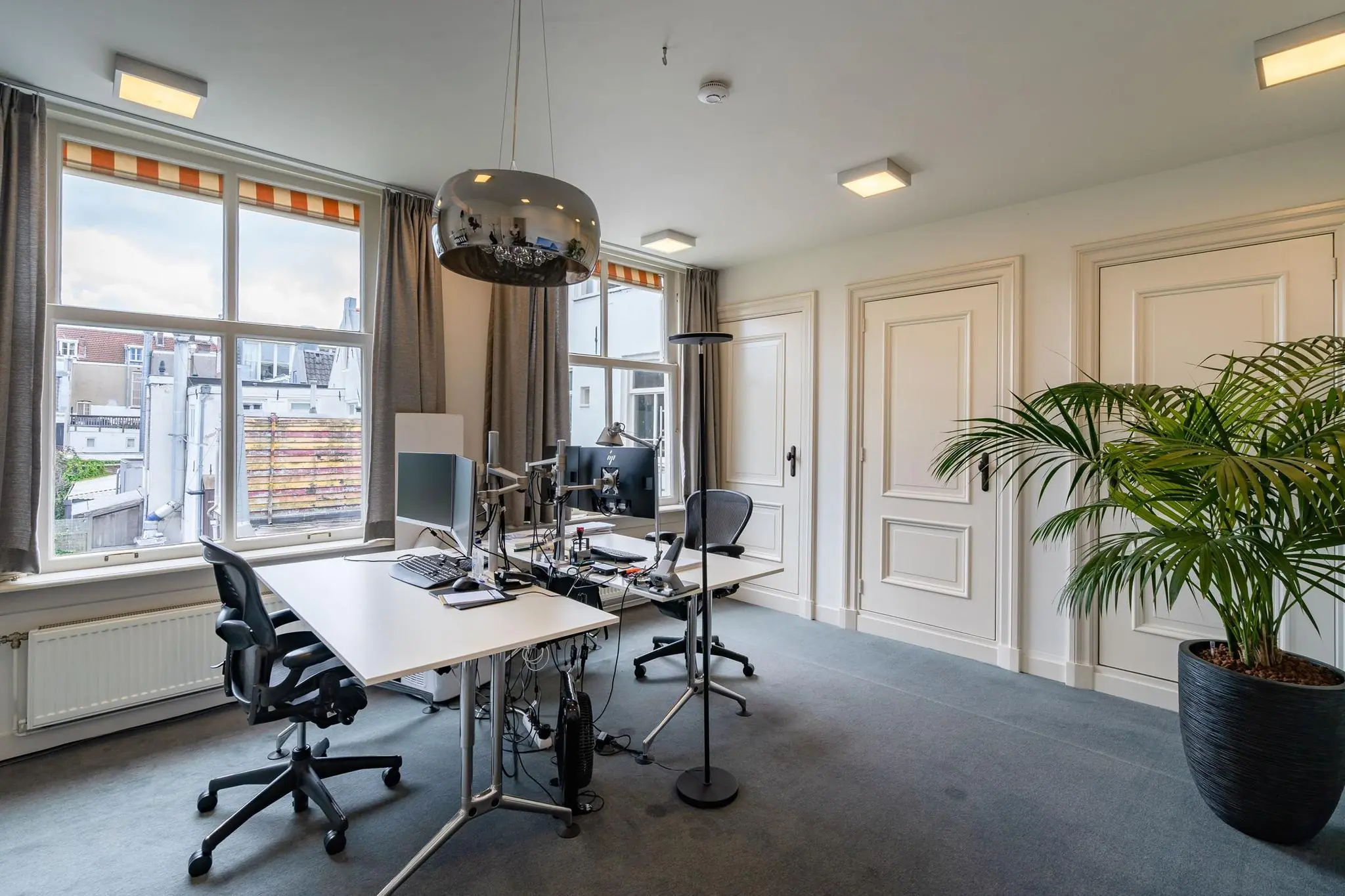 Modern kantoorinterieur aan de Herengracht met twee werkplekken, grote ramen en een groene kamerplant.