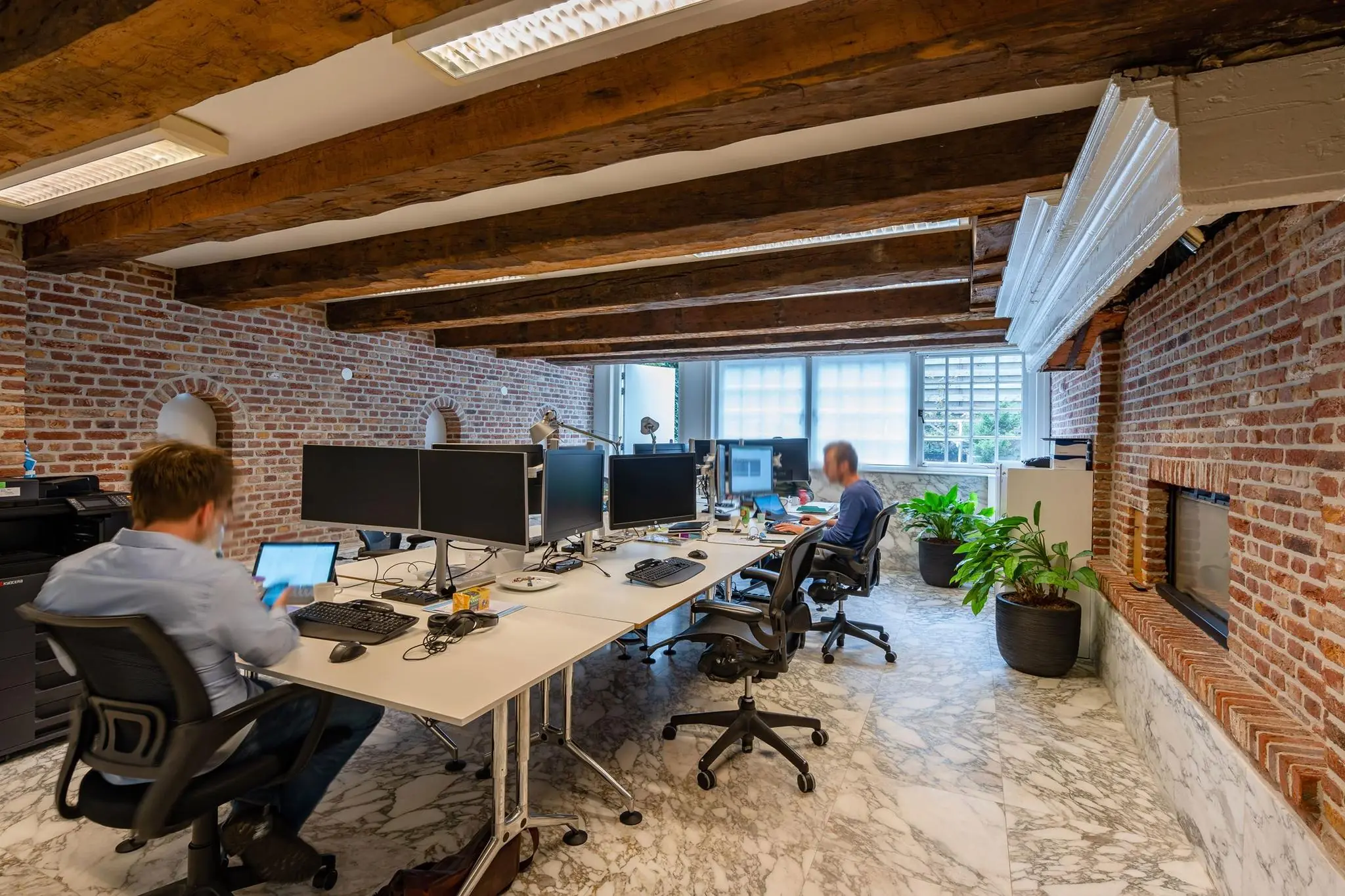 Moderne kantoorruimte aan de Herengracht met zichtbare bakstenen muren, houten plafondbalken en werkplekken met meerdere beeldschermen.