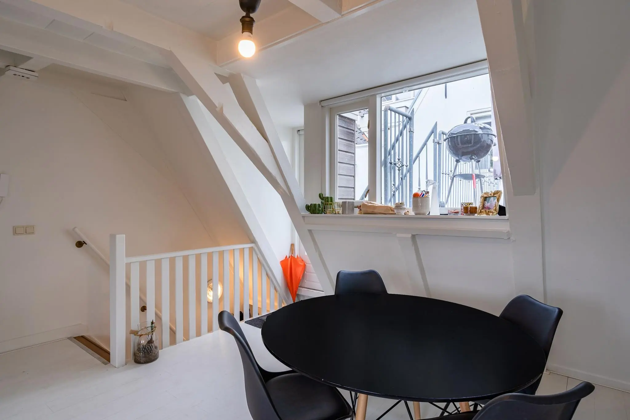 Witgeschilderde zolderkamer aan de Herengracht met een zwarte eettafel, zicht op een buitentrap en barbecue door het raam.
