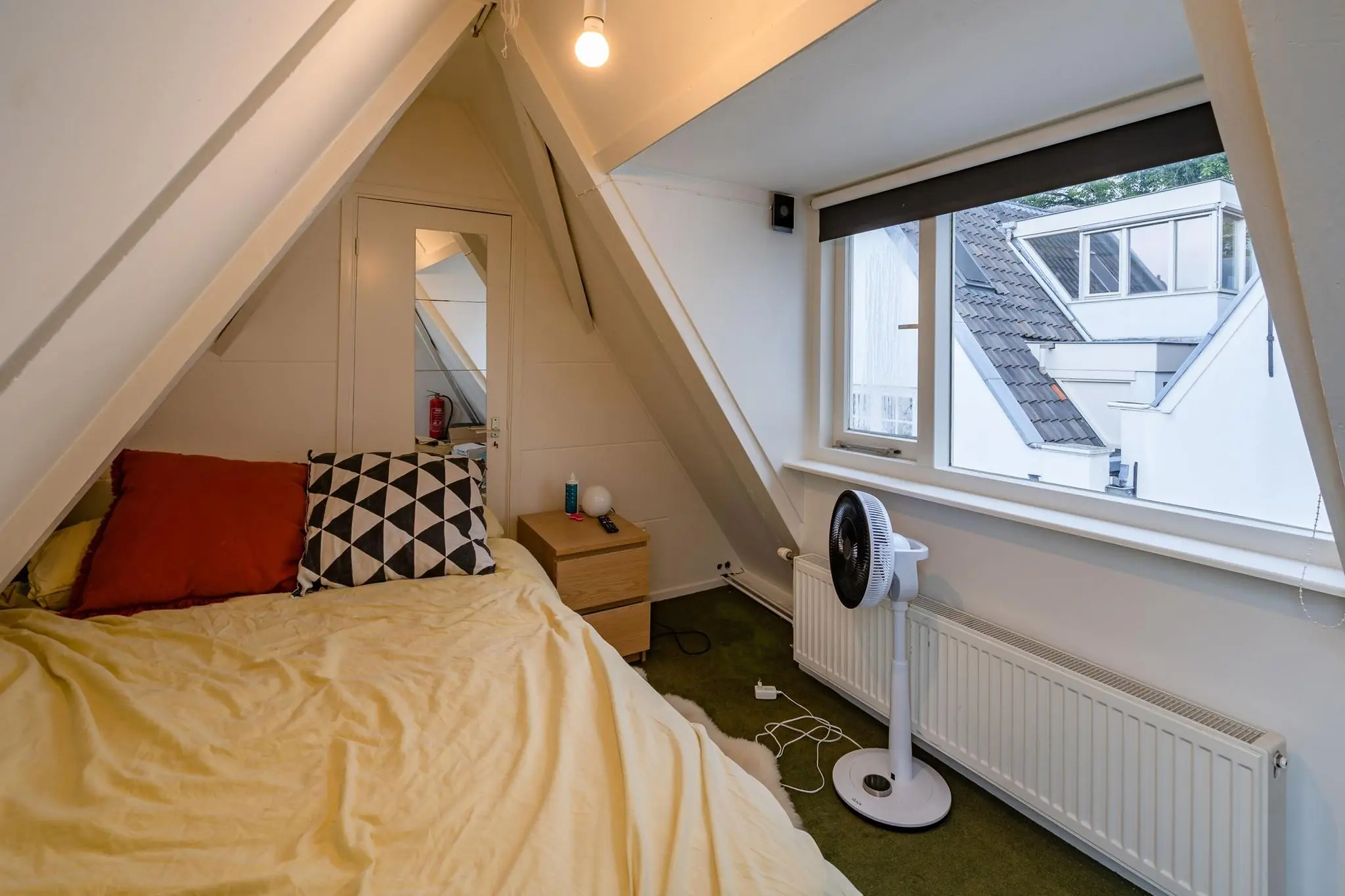 Kleine zolderkamer aan de Herengracht met een schuin dak, een eenpersoonsbed, ventilator en uitzicht op witte daken.