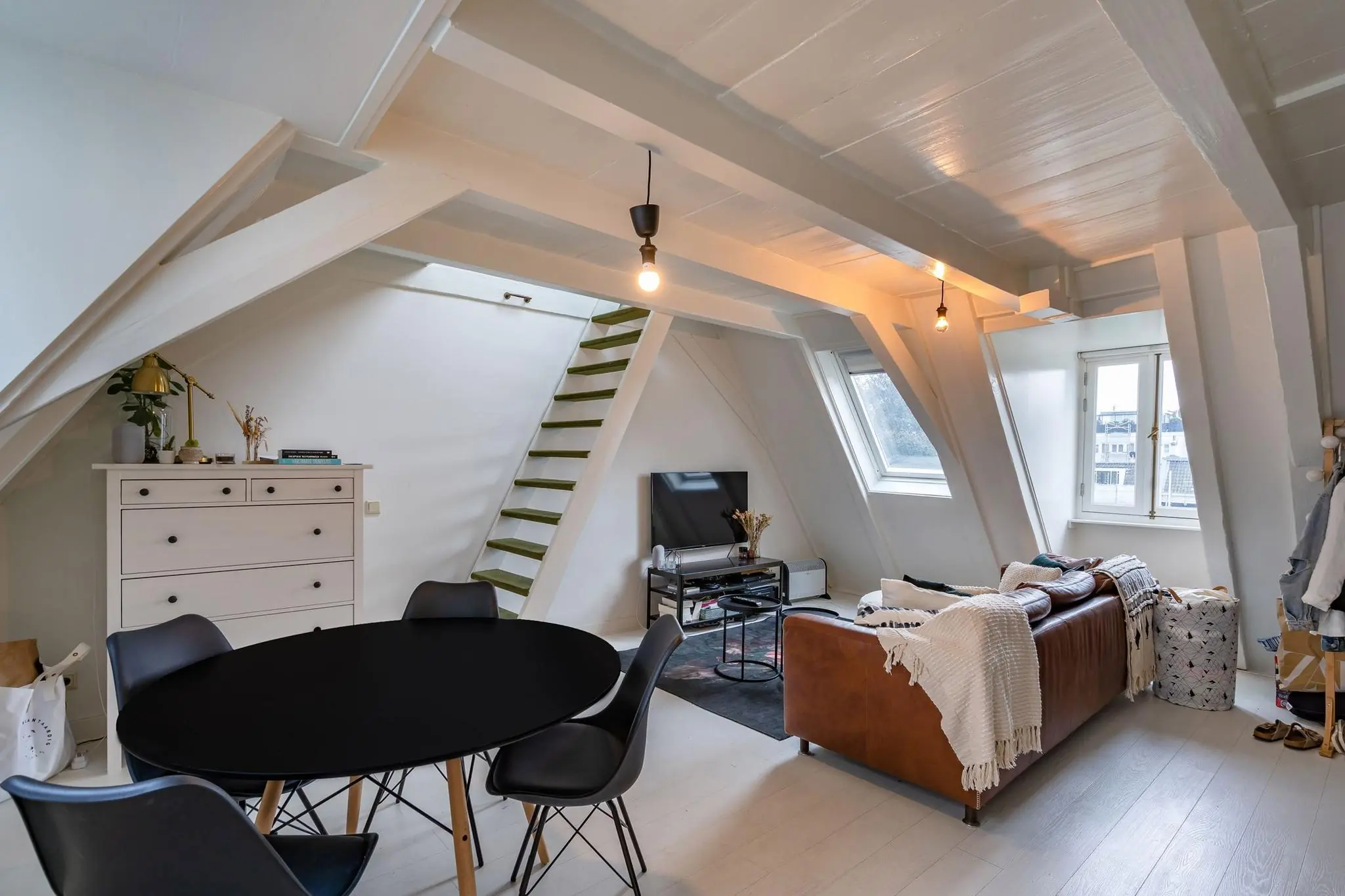 Licht ingerichte zolderwoonkamer aan de Herengracht met een leren bank, zwarte eettafel en een trap naar een hoger gelegen slaapgedeelte.