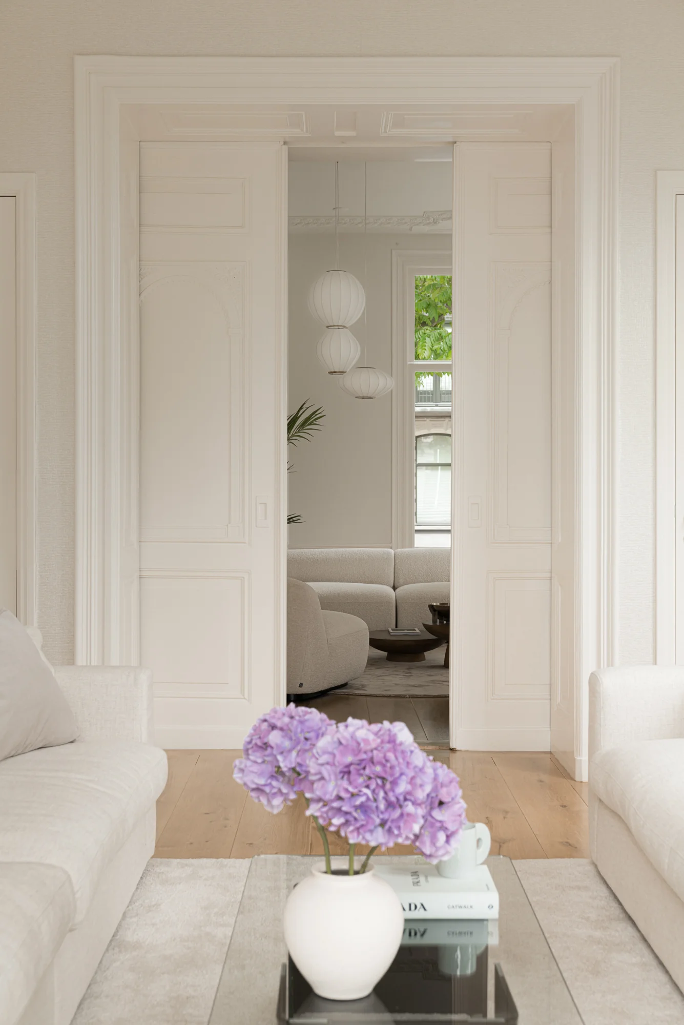 Licht ingerichte woonkamer aan de Weteringschans met paarse bloemen op een salontafel en zicht op een tweede zithoek door schuifdeuren.