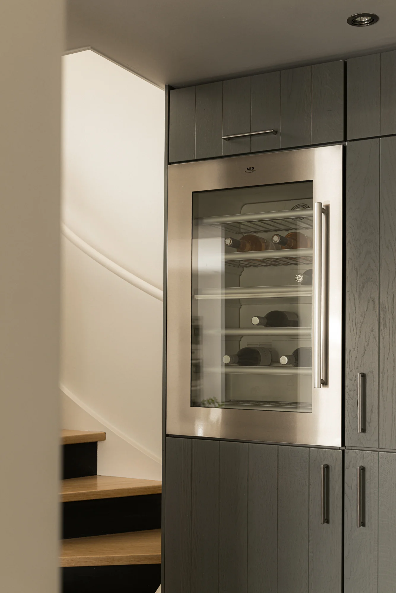 Ingebouwde wijnklimaatkast van AEG in een moderne donkere keukenkast naast een houten trap.