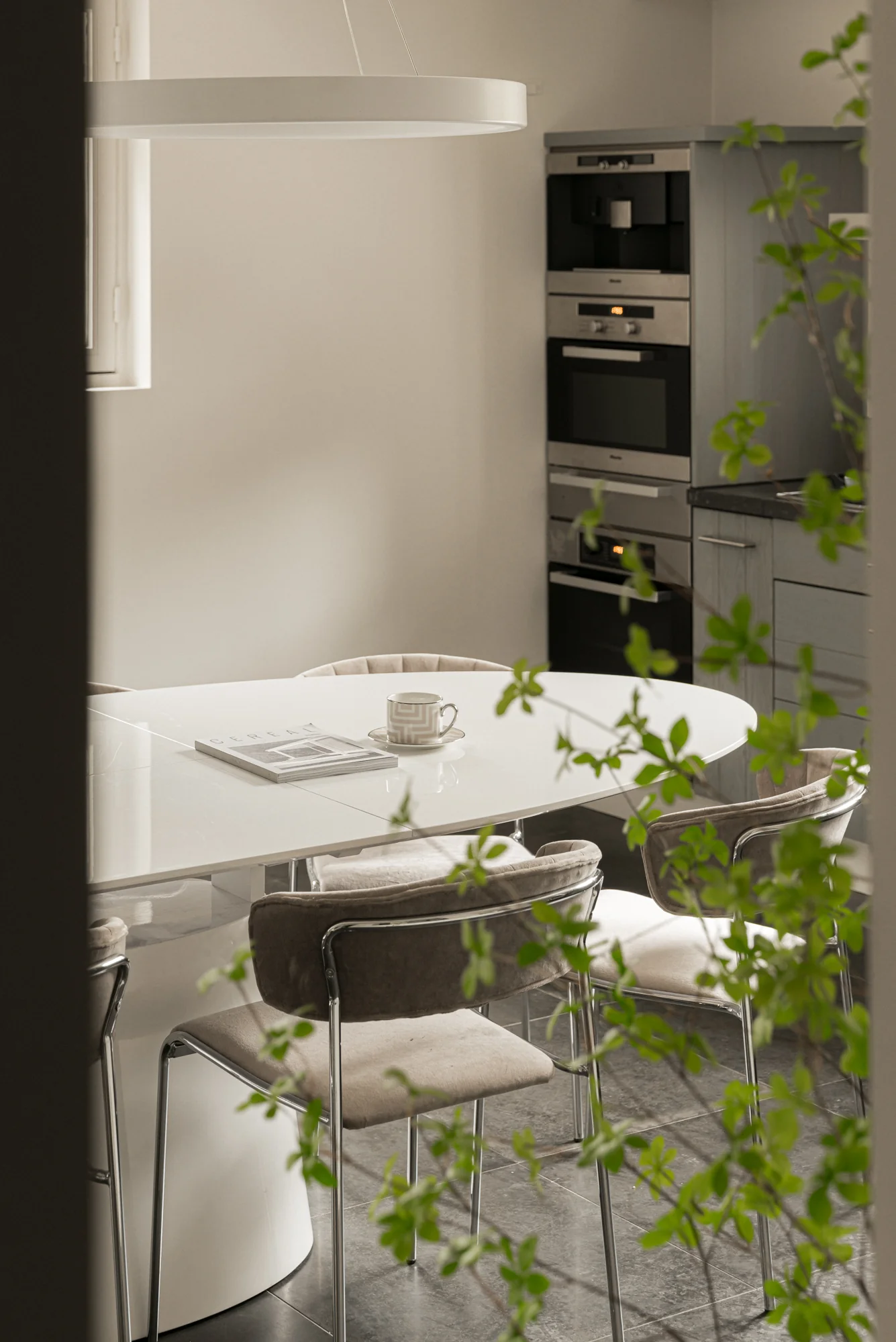 Moderne keuken met een witte eettafel, omringd door zachte stoelen en natuurlijke lichtinval, gezien vanaf een deuropening met groen.