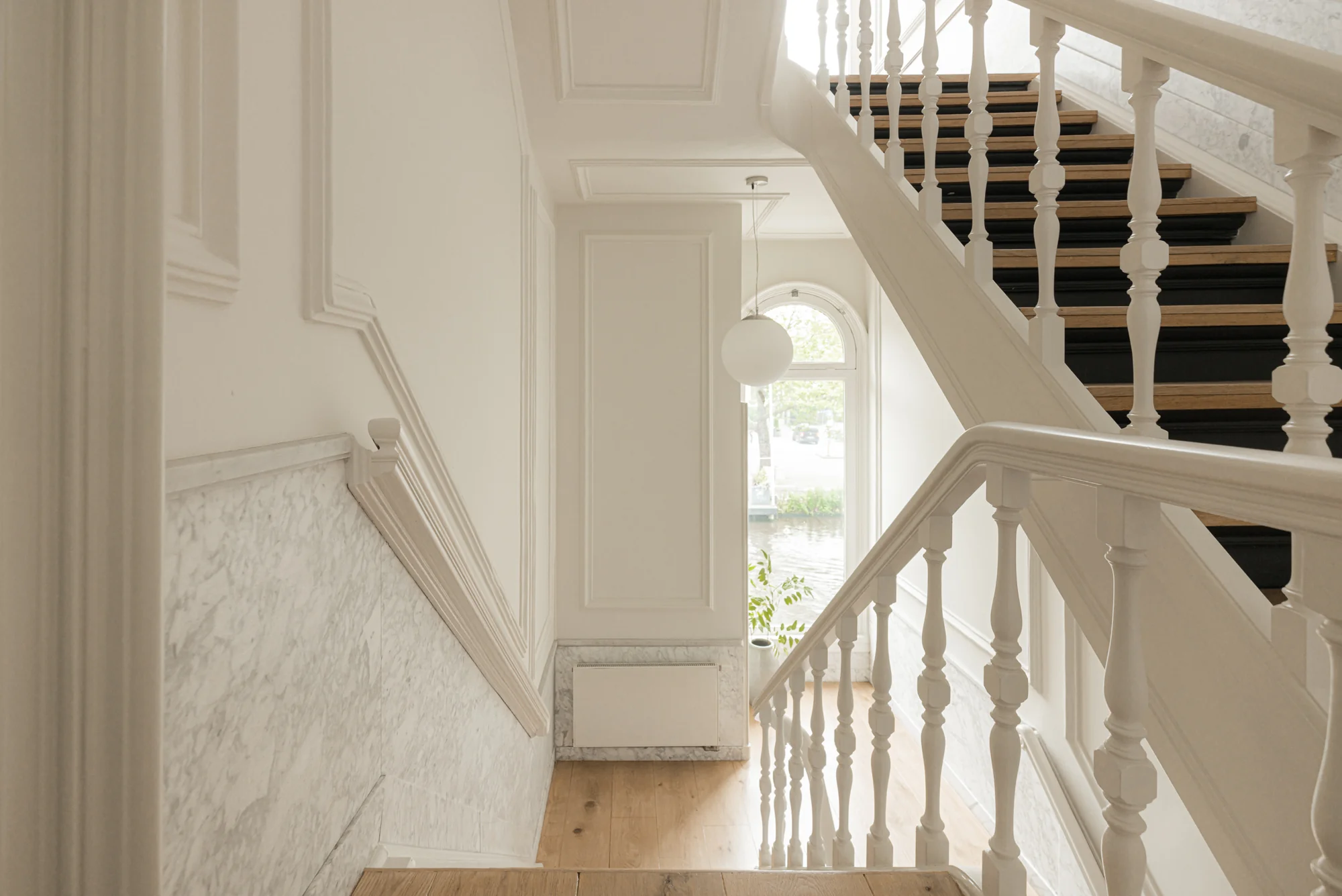 Licht trappenhuis aan de Weteringschans met witte balustrades, houten treden en een groot raam met zicht op de gracht.