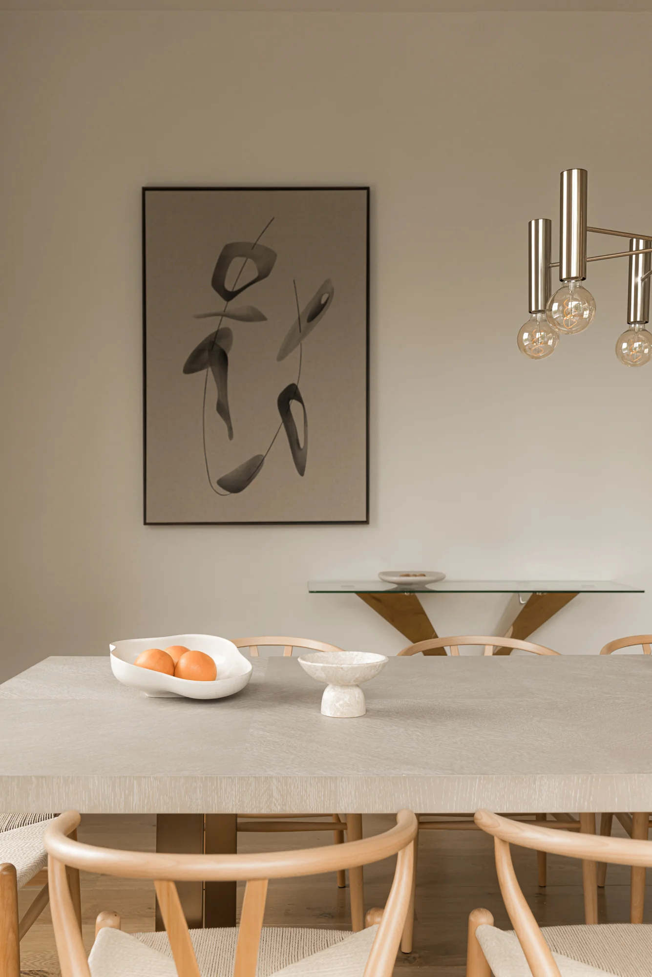 Minimalistisch ingerichte eethoek met houten stoelen, een lichtgrijze tafel, modern wandkunstwerk en designverlichting aan het plafond.
