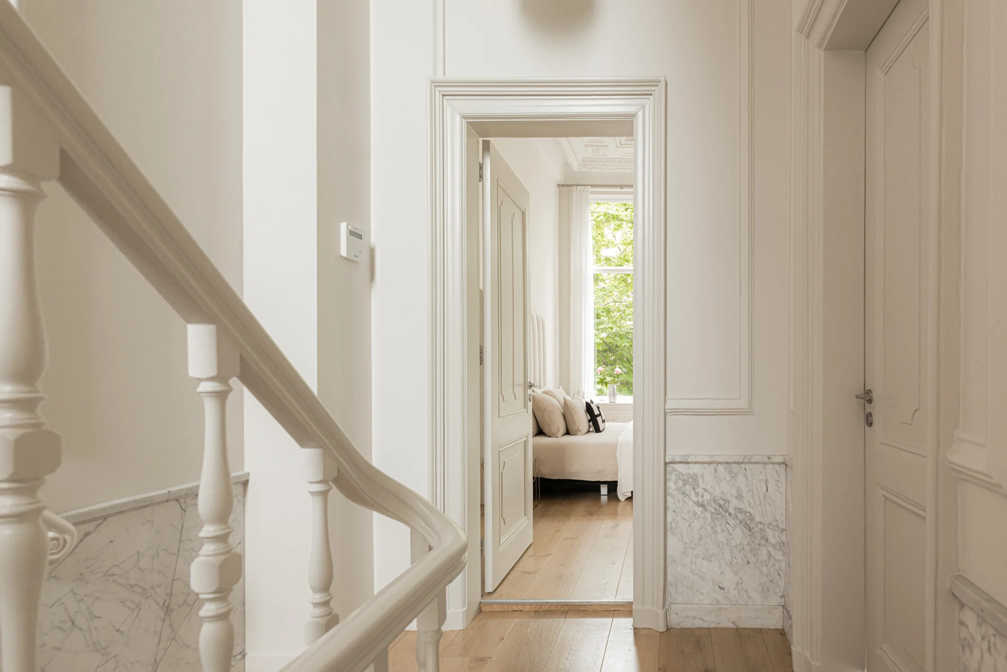 Lichte en stijlvolle hal met houten vloer, witte trapleuning en doorkijk naar een slaapkamer met grote raam.