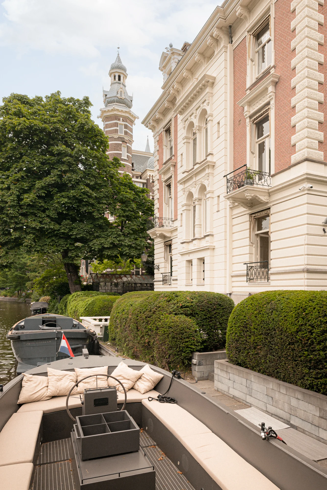 Luxe boot met zitkussens ligt aangemeerd aan de gracht bij statige panden aan de Weteringschans in Amsterdam.