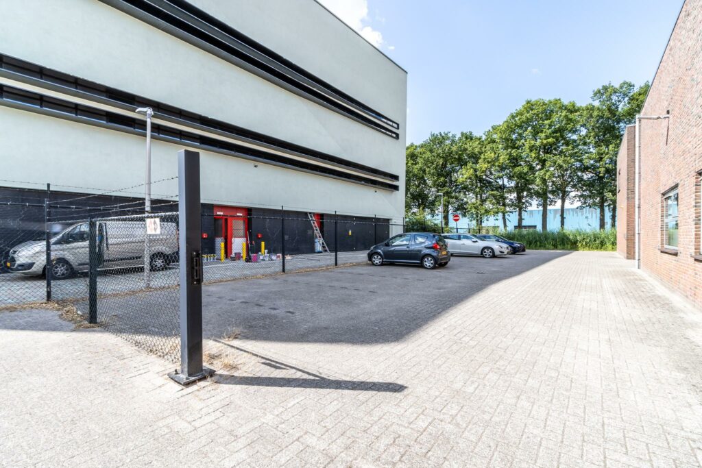 Parkeerterrein aan de Terminalweg met geparkeerde auto's naast een modern bedrijfsgebouw en een omheind terrein.