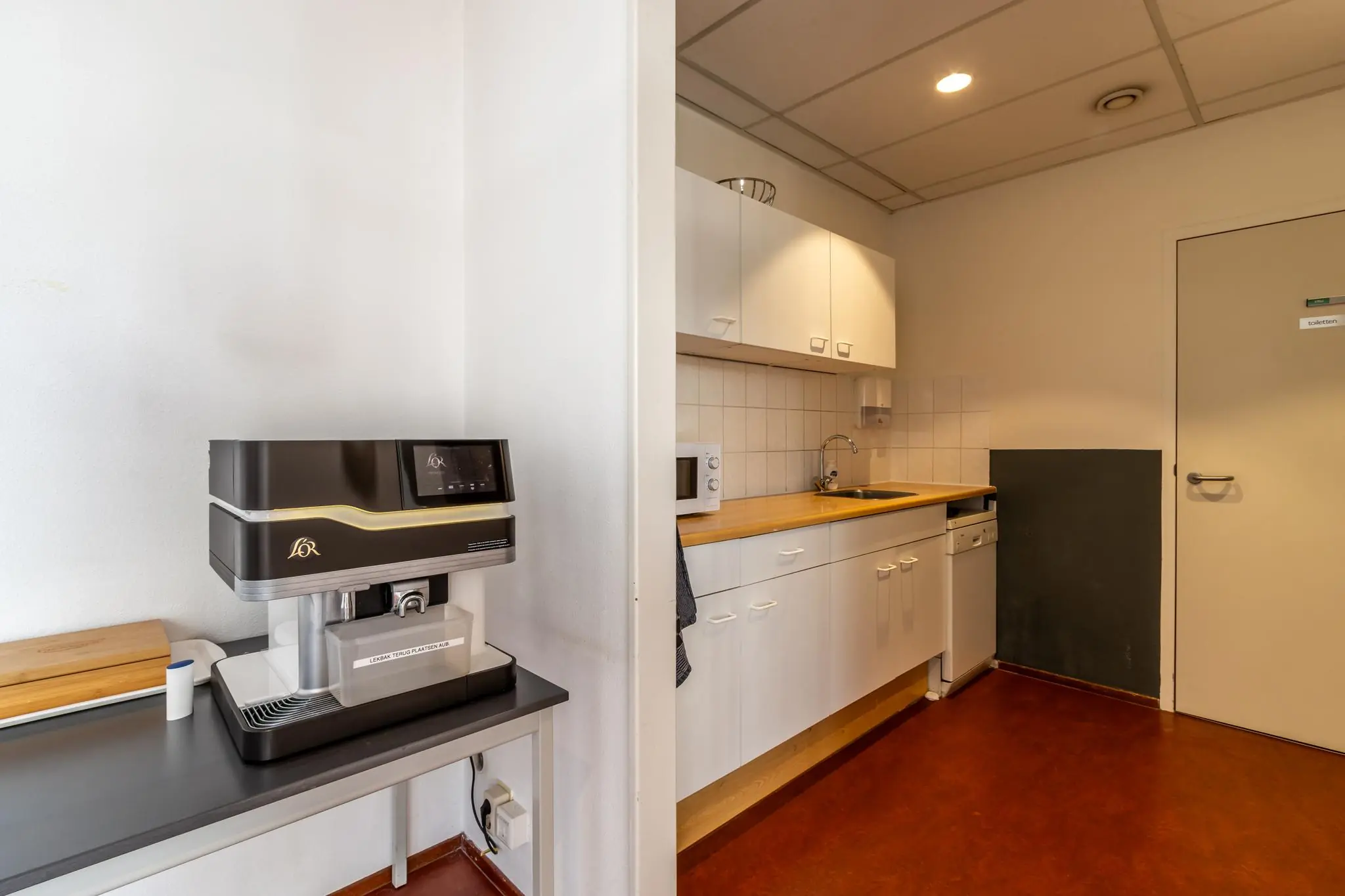Kleine kantoorkeuken met een koffieautomaat, aanrecht, magnetron en een deur naar het toilet.