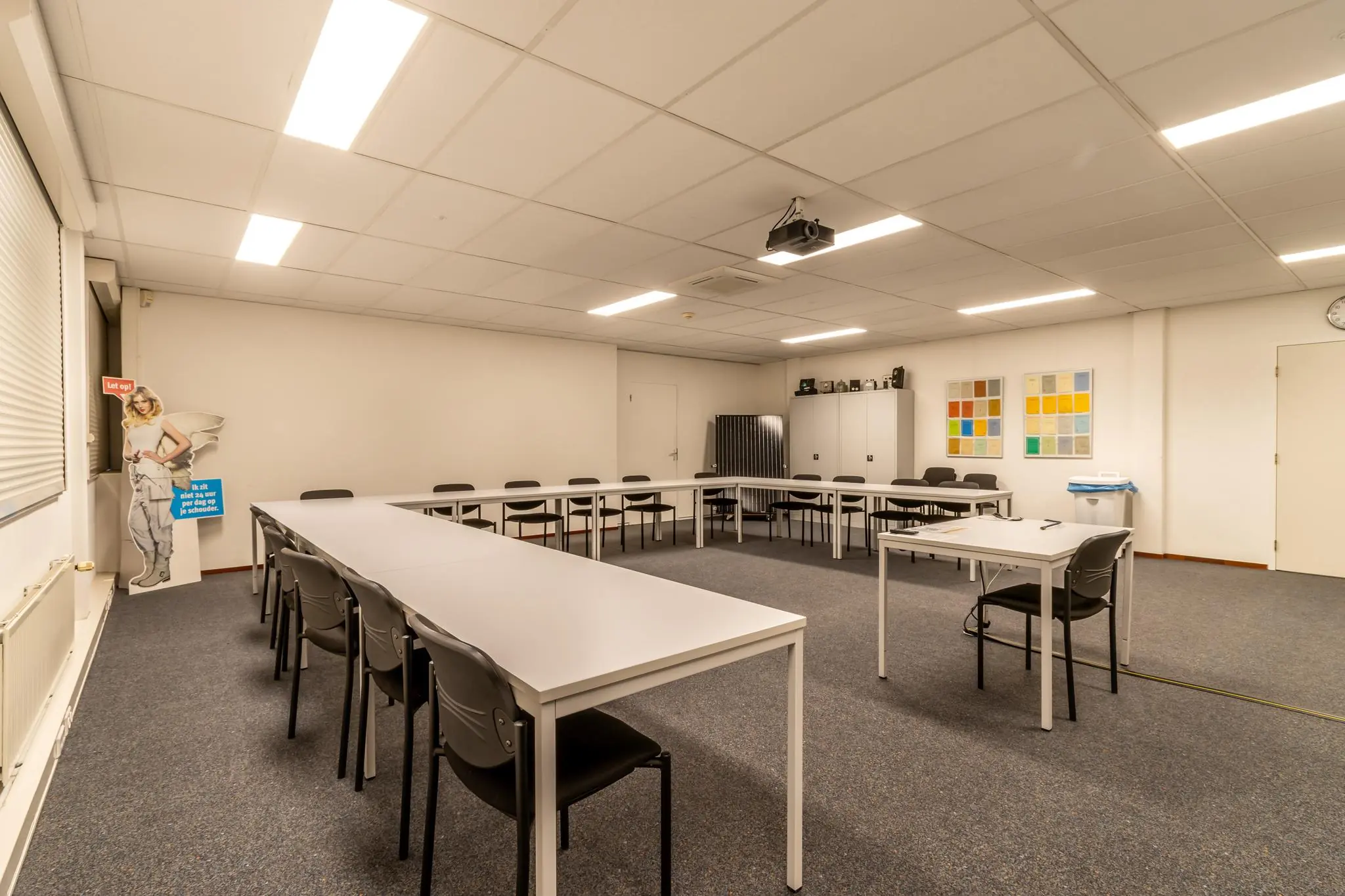 Vergaderruimte aan de Terminalweg met U-vormige opstelling van tafels, stoelen, projector en een kartonnen waarschuwingsbord in de hoek.