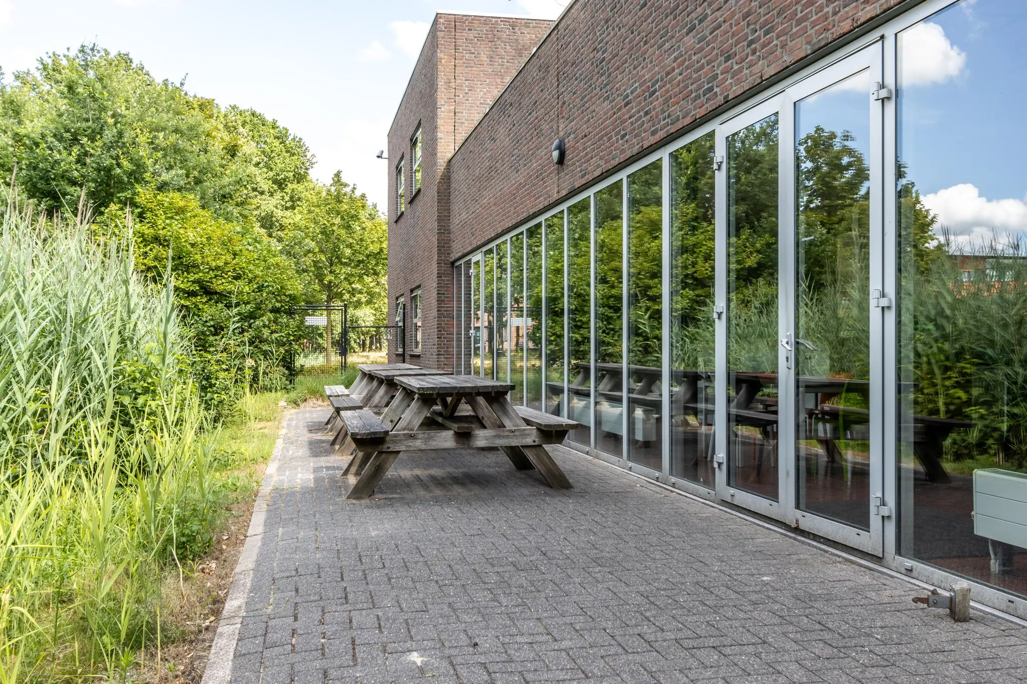Houten picknicktafels staan langs de glazen gevel van een modern bakstenen gebouw aan de Terminalweg, omringd door groen.