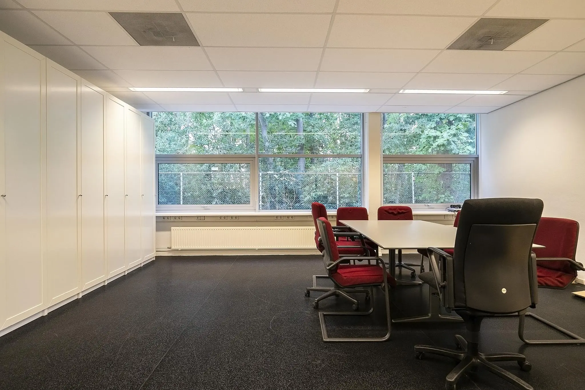 Vergaderruimte aan de Banningstraat met een lange tafel, rode stoelen, grote ramen en ingebouwde kasten.