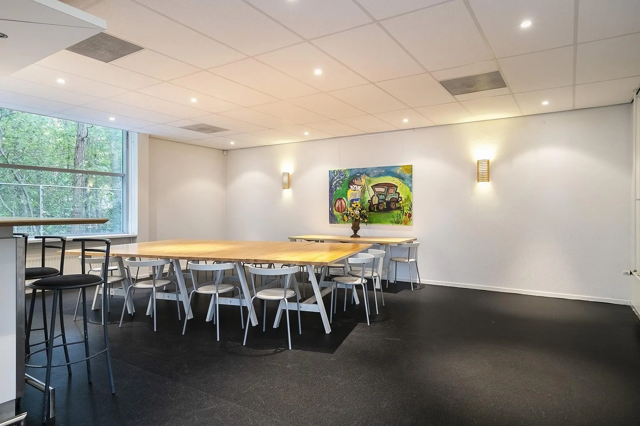 Vergaderruimte met grote houten tafel, witte stoelen, kleurrijk schilderij en een groot raam met uitzicht op bomen.
