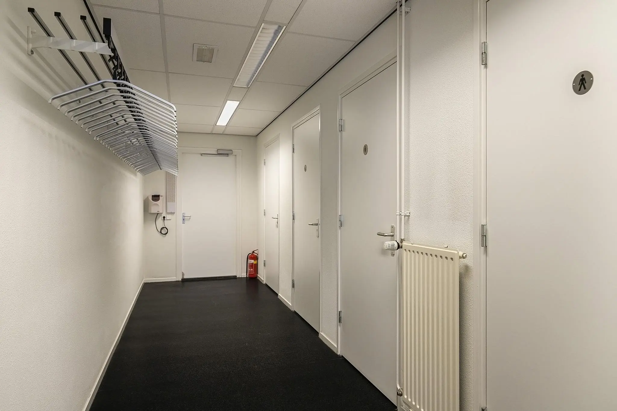 Witte gang met meerdere deuren, een kapstok met hangers en een brandblusser in een gebouw aan de Banningstraat.