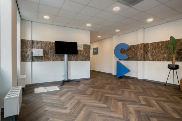 Moderne ontvangsthal met houten visgraatvloer, een groot blauw logo, tv-scherm en plantenbak.