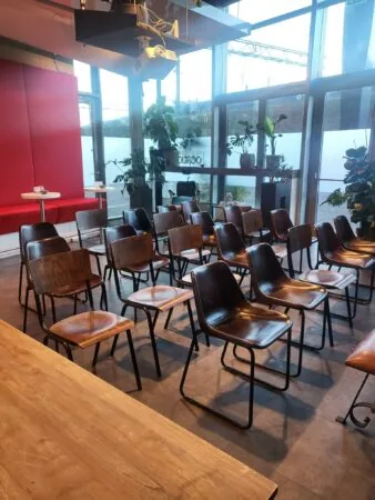 Kleine vergaderruimte aan de Oosterdoksstraat met houten stoelen, rode wandbekleding en grote ramen met uitzicht naar buiten.