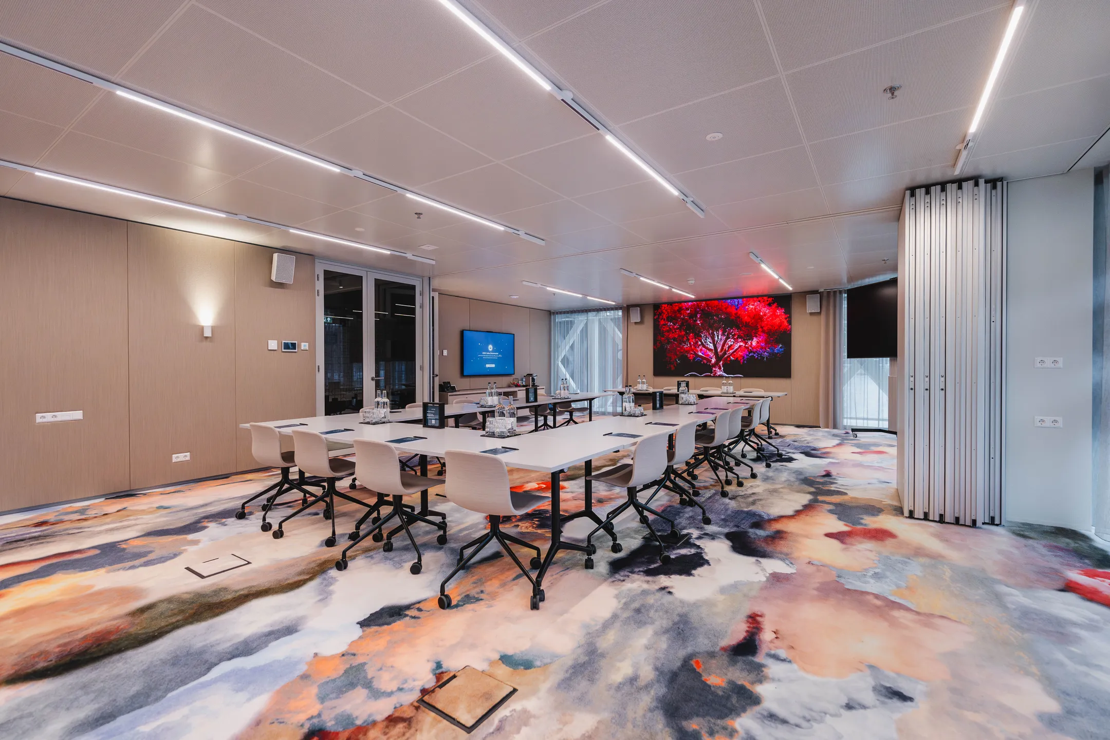 Moderne vergaderruimte met kleurrijk tapijt, schermen en een kunstwerk van een rode boom.