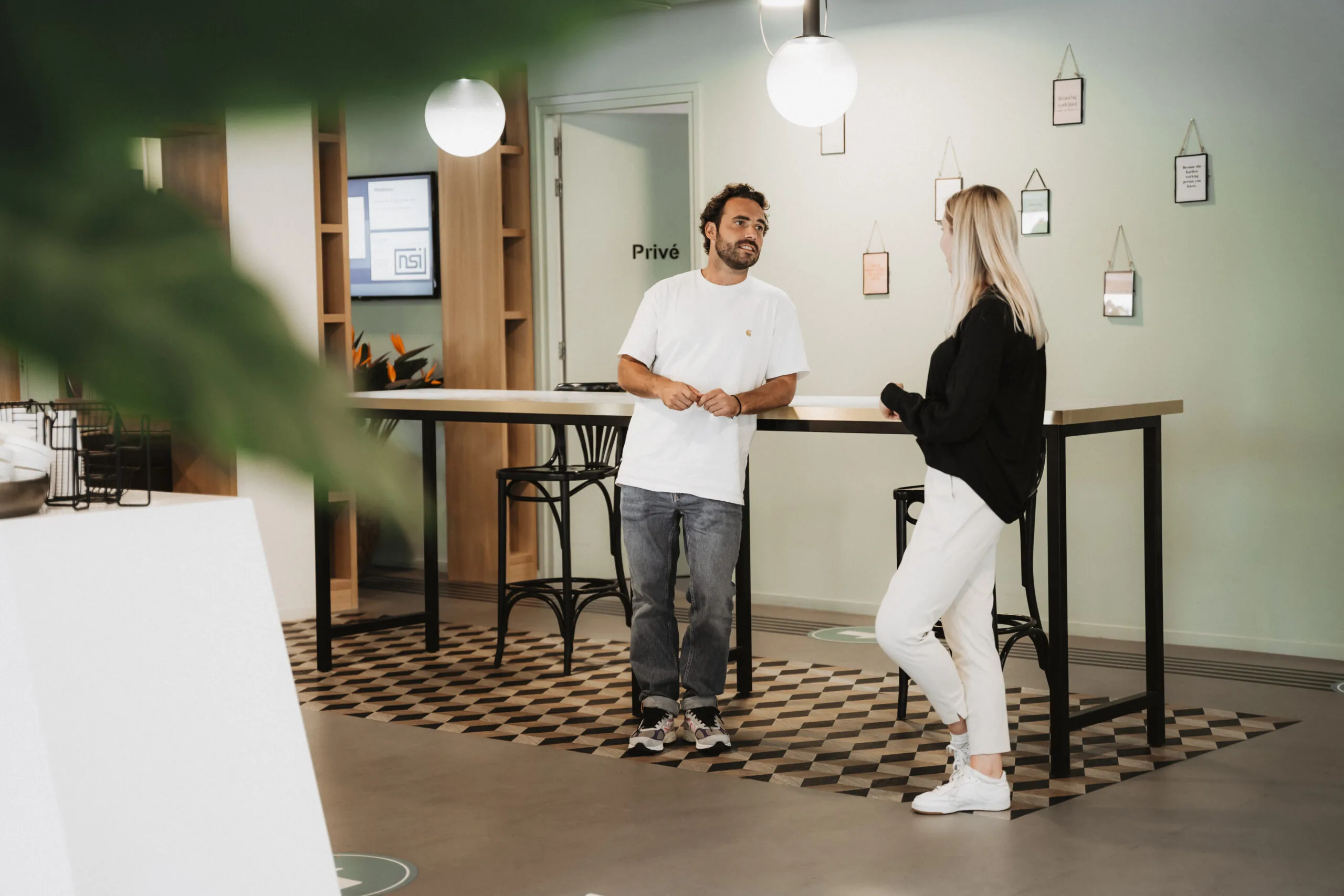 Twee mensen praten informeel met elkaar bij een hoge tafel in de moderne ontvangsthal van HNK Amsterdam Schinkel.