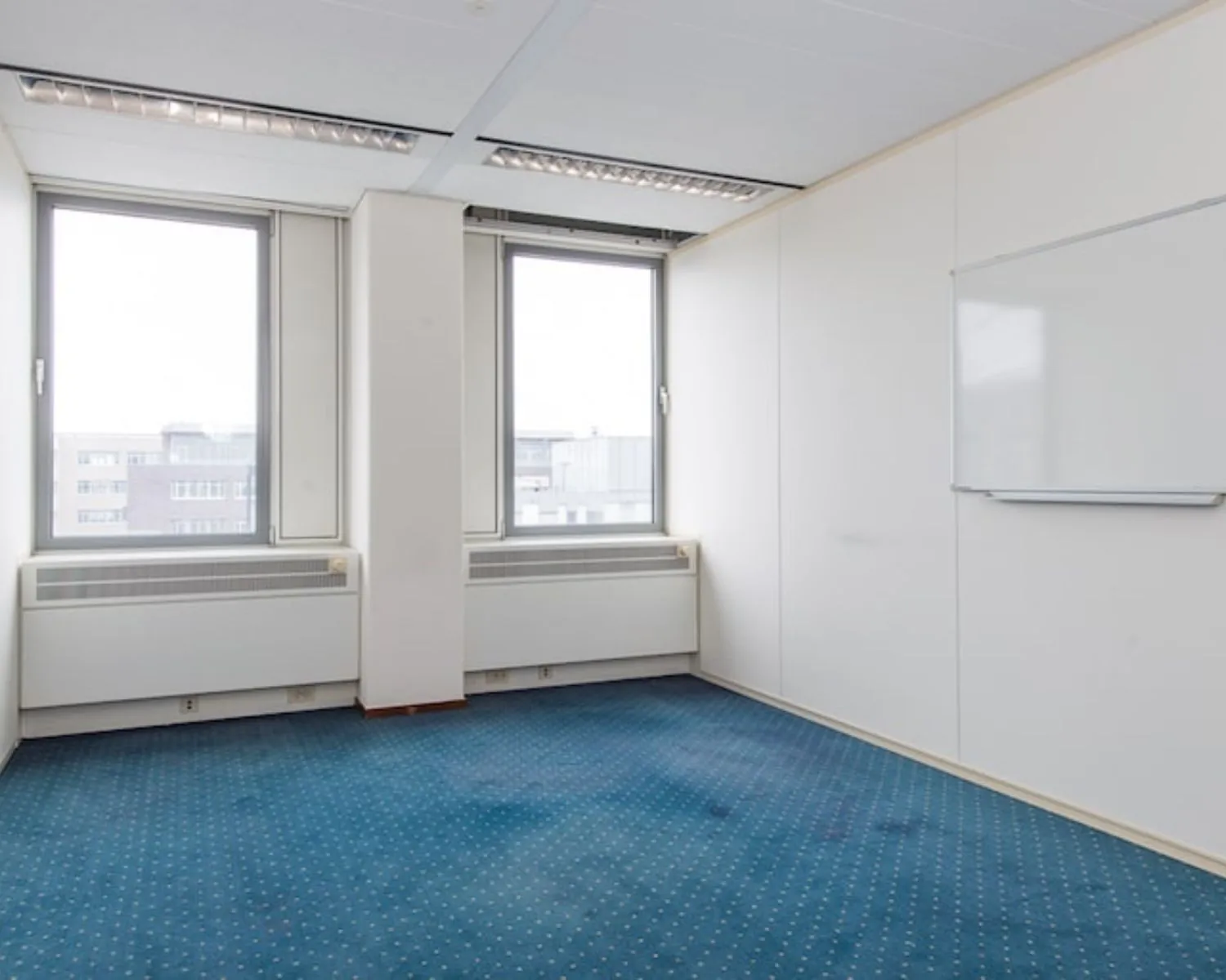 Lichte kantoorruimte met blauw tapijt, twee grote ramen en een whiteboard aan de muur aan de Gaetano Martinolaan 75-95 in Maastricht.