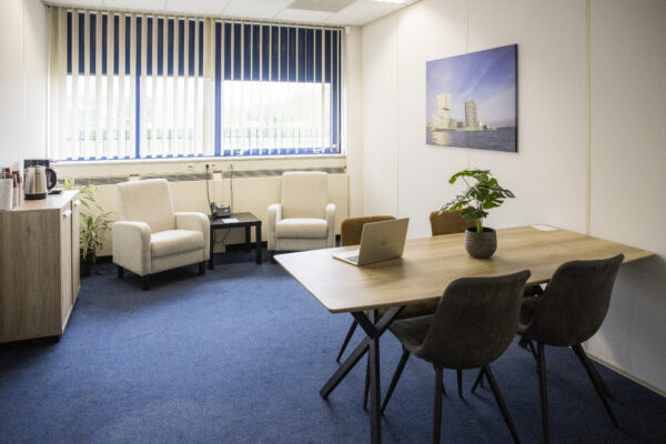 Een licht kantoor met een tafel, stoelen, twee fauteuils, een laptop, en een foto aan de muur.