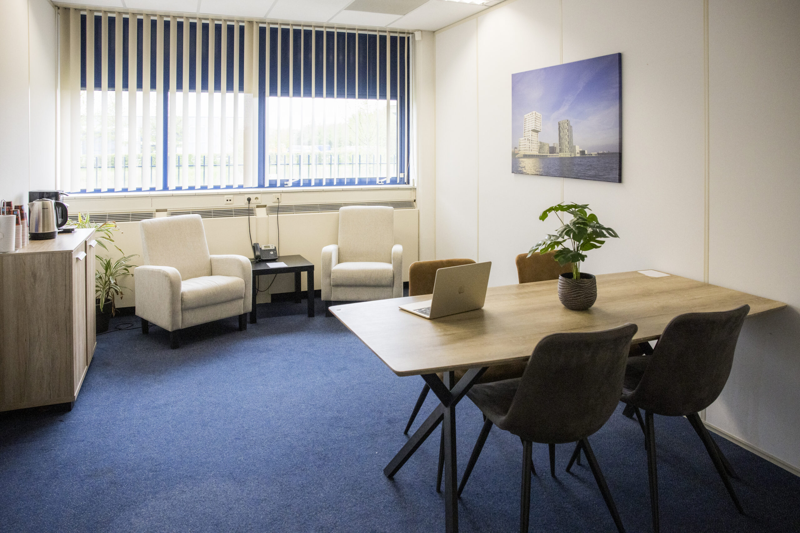 Een licht kantoor met een tafel, stoelen, twee fauteuils, een laptop, en een foto aan de muur.