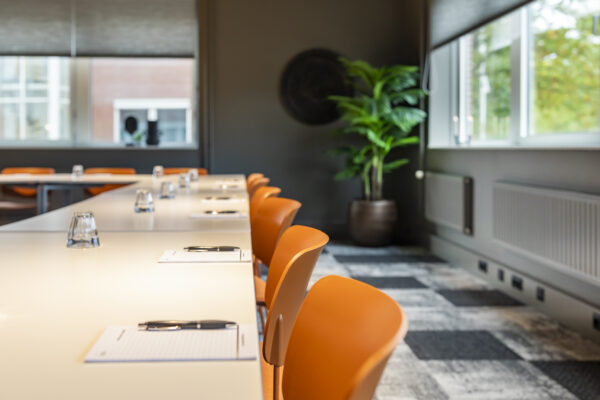 Een vergaderruimte met oranje stoelen en een tafel met schrijfblokken en pennen.