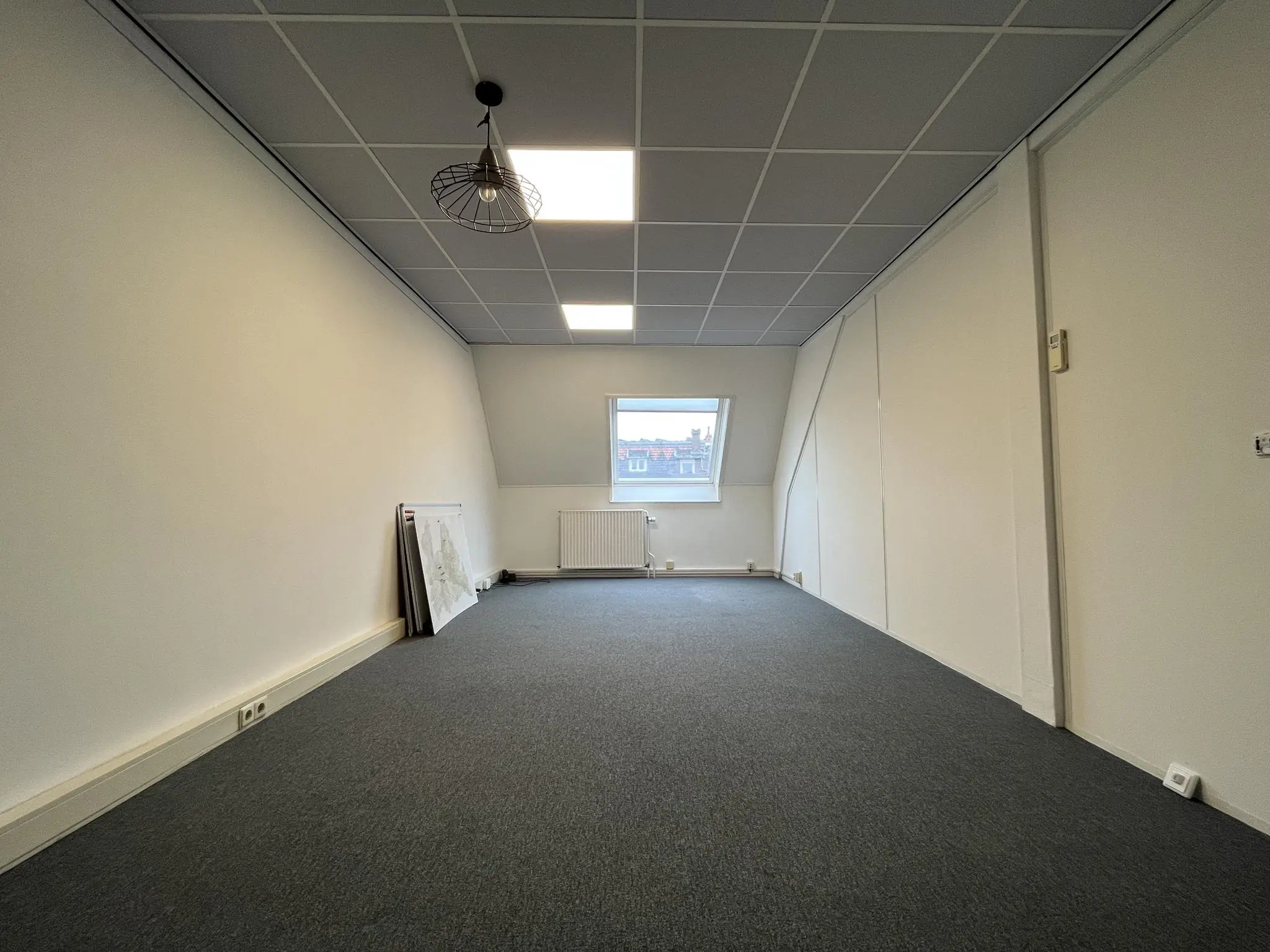 Leeg kantoorruimte aan de Ramstraat met schuin plafond, systeemplafond, enkel raam en enkele op de vloer geleunde kaarten of posters.