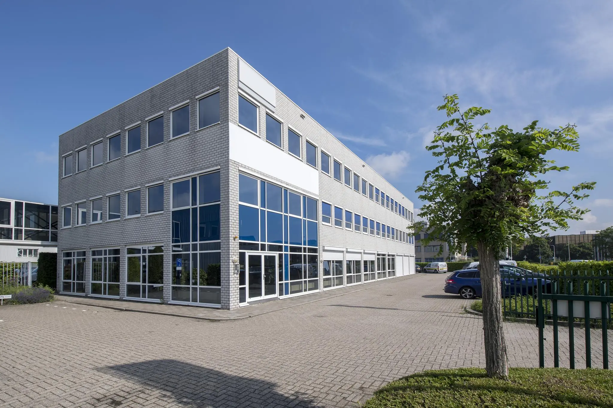 Modern kantoorgebouw met grote ramen en een parkeerterrein op een zonnige dag.