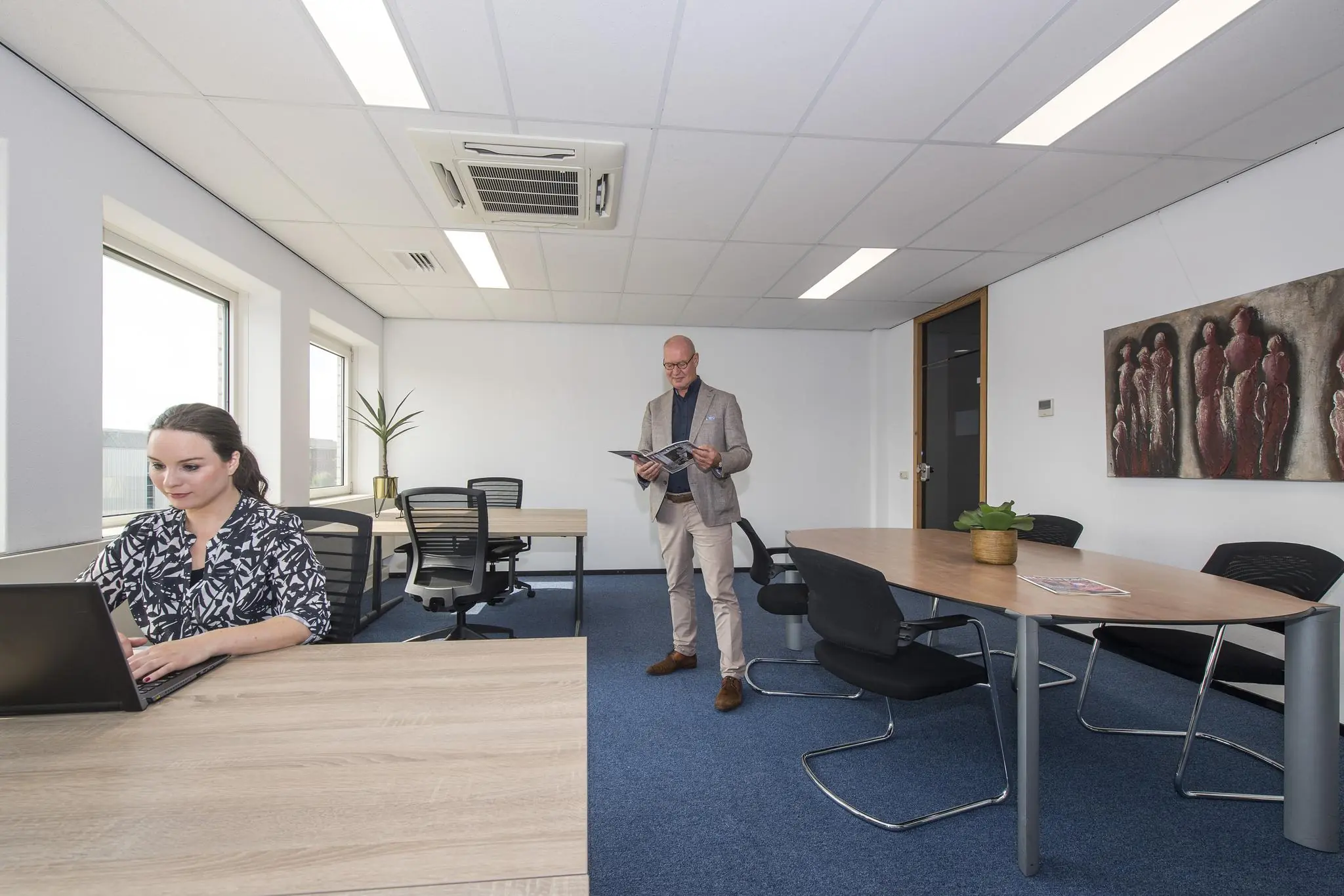 Twee mensen aan het werk in een modern kantoor met houten bureaus en blauwe vloerbedekking.