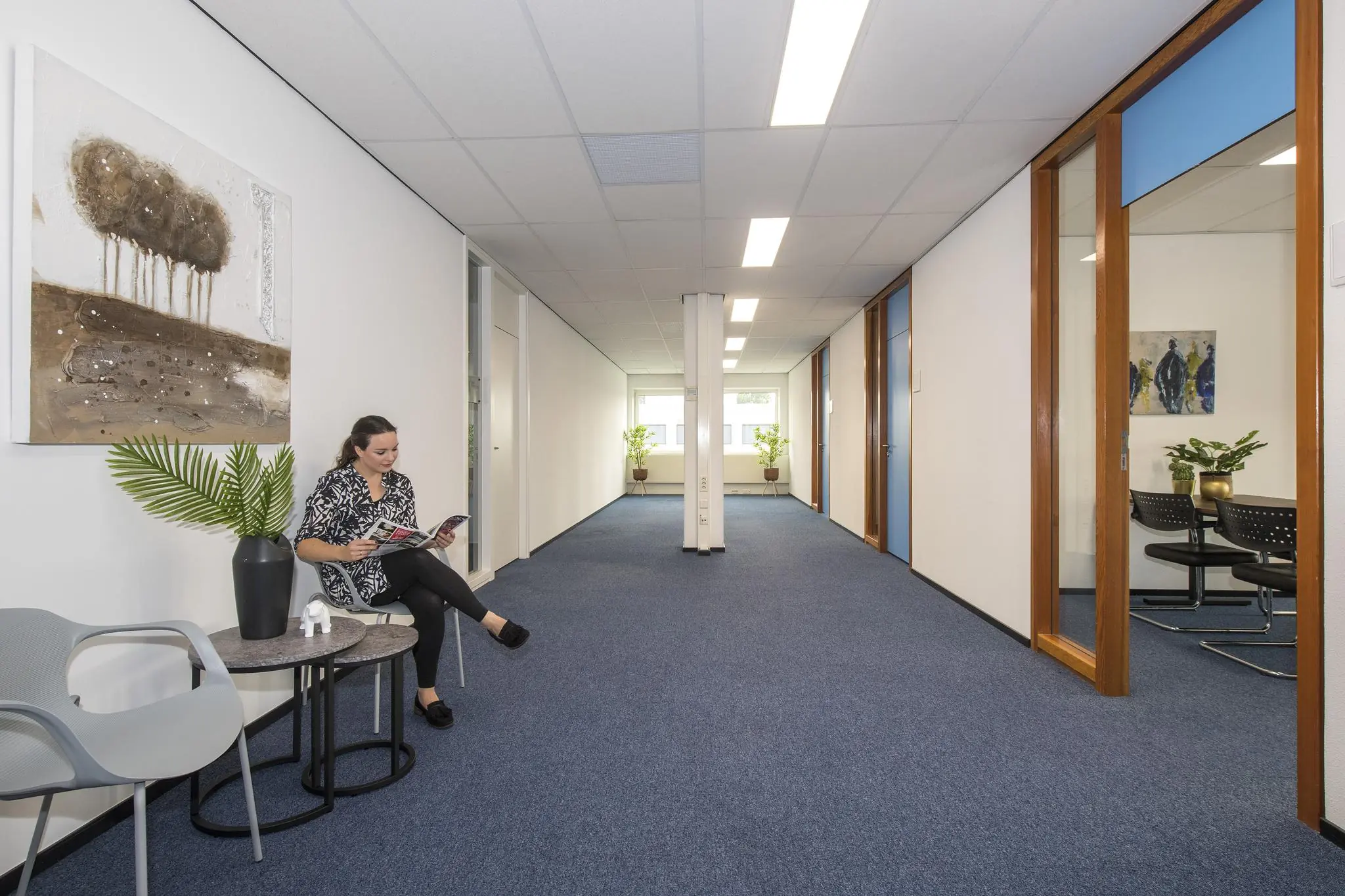 Vrouw leest een tijdschrift in een wachtruimte van een kantoor, met het schilderij 'Witboom' aan de muur.