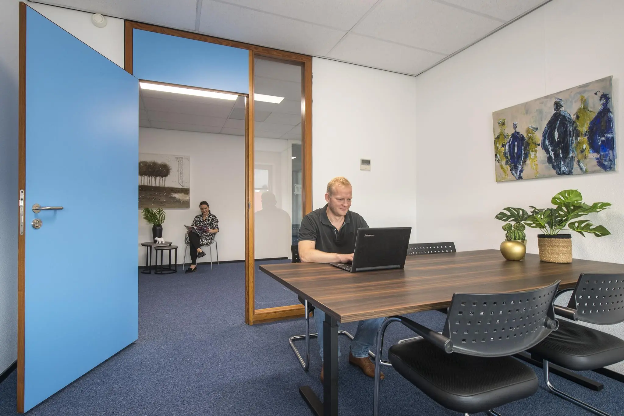 Man werkt op laptop aan vergadertafel in kantoorruimte met open deur naar wachtruimte waar een vrouw een tijdschrift leest.