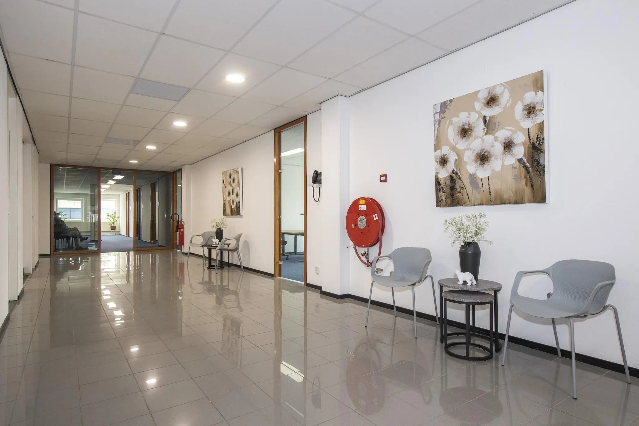 Wachtgebied in een kantoorpand met grijze stoelen, bloemenkunst aan de muur en een brandslanghaspel.