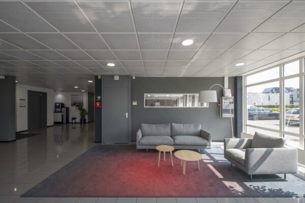 Moderne wachtruimte met grijze zitbanken, houten tafeltjes en grote ramen die uitzicht bieden op een bedrijventerrein.
